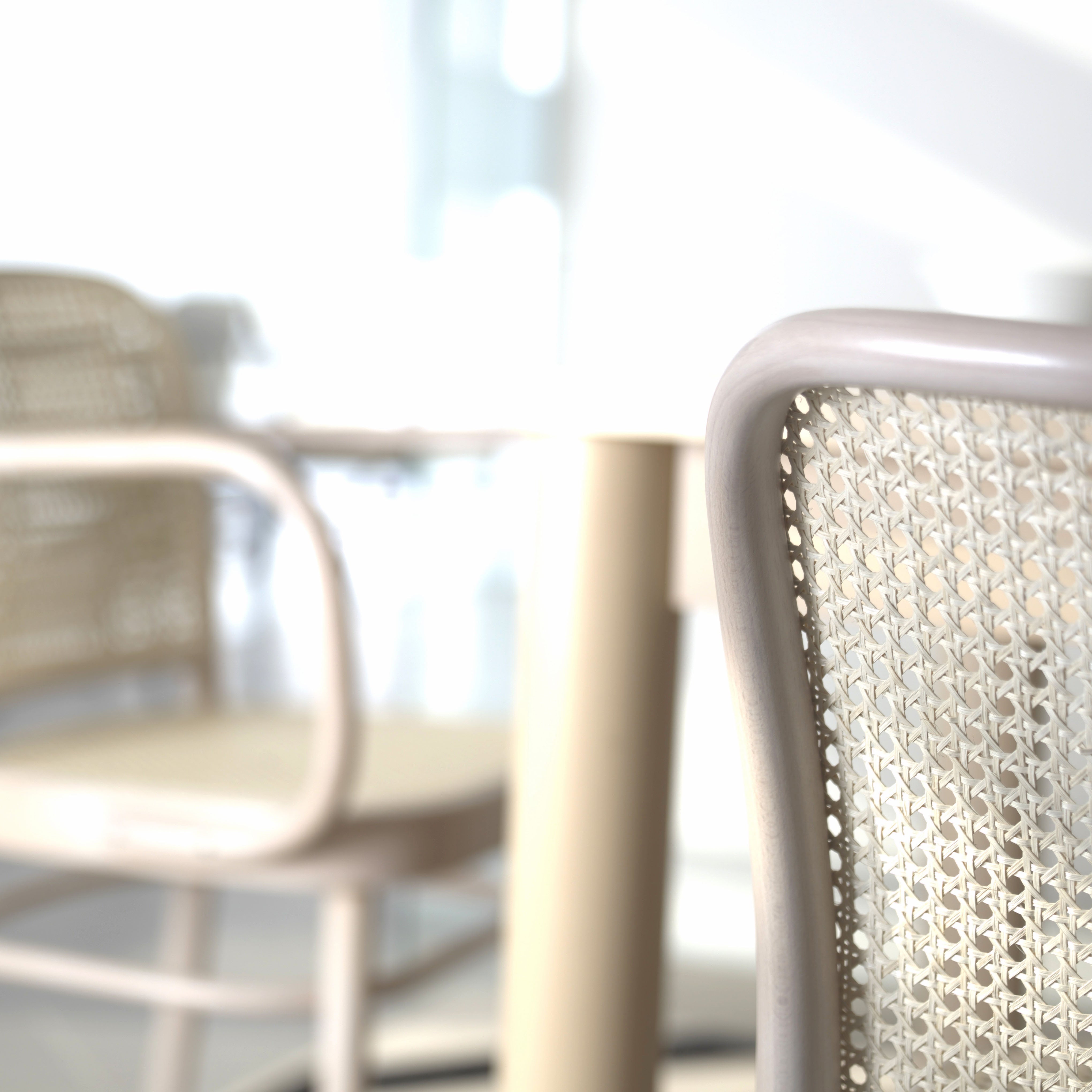 Close-up of a Chair 811 - Cane by TON in a sunlit room, reflecting Art Nouveau elegance. A blurred background features another Josef Hoffmann design with a round table, casting soft shadows on the light floor, creating a bright and airy atmosphere.