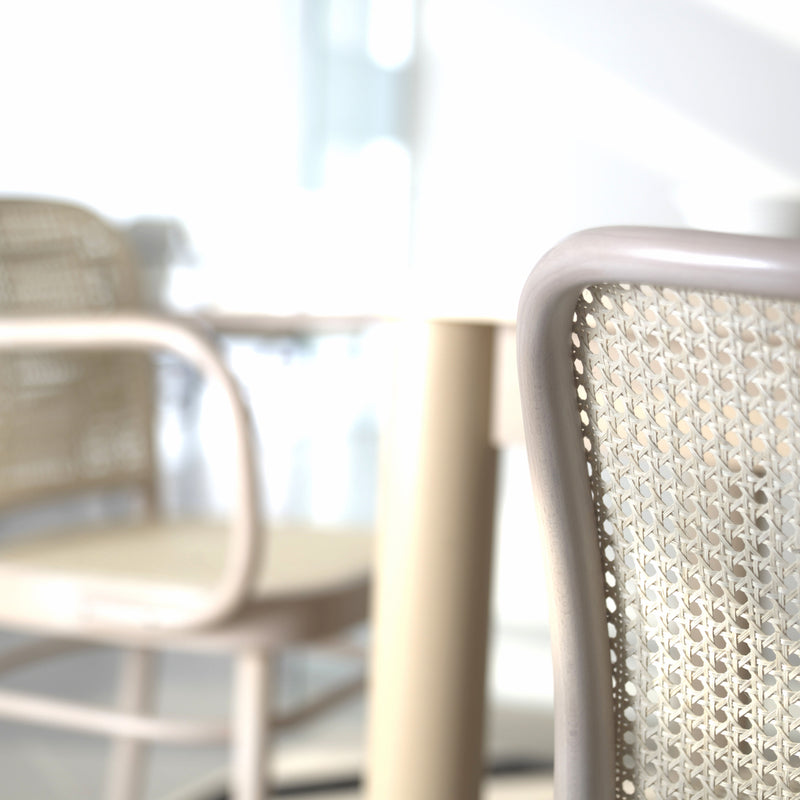 Close-up of a Chair 811 - Cane by TON in a sunlit room, reflecting Art Nouveau elegance. A blurred background features another Josef Hoffmann design with a round table, casting soft shadows on the light floor, creating a bright and airy atmosphere.
