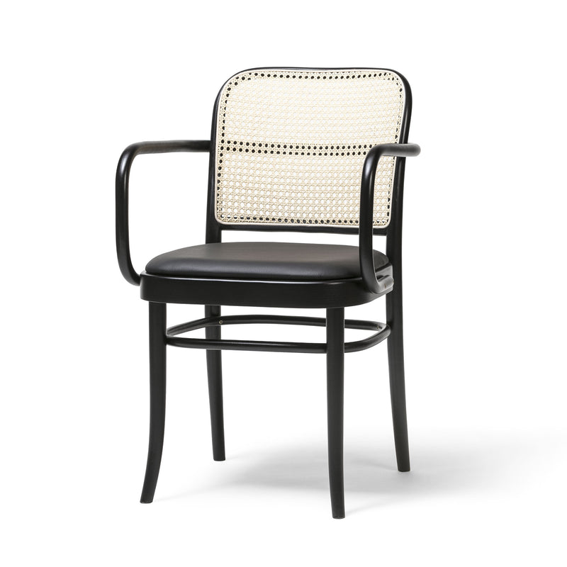 The Armchair 811 by TON, a black wooden chair with a woven cane backrest and cushioned seat, embodies Josef Hoffmanns elegant simplicity. Its curved armrests and sleek lines evoke Art Nouveau aesthetics against a plain white backdrop.