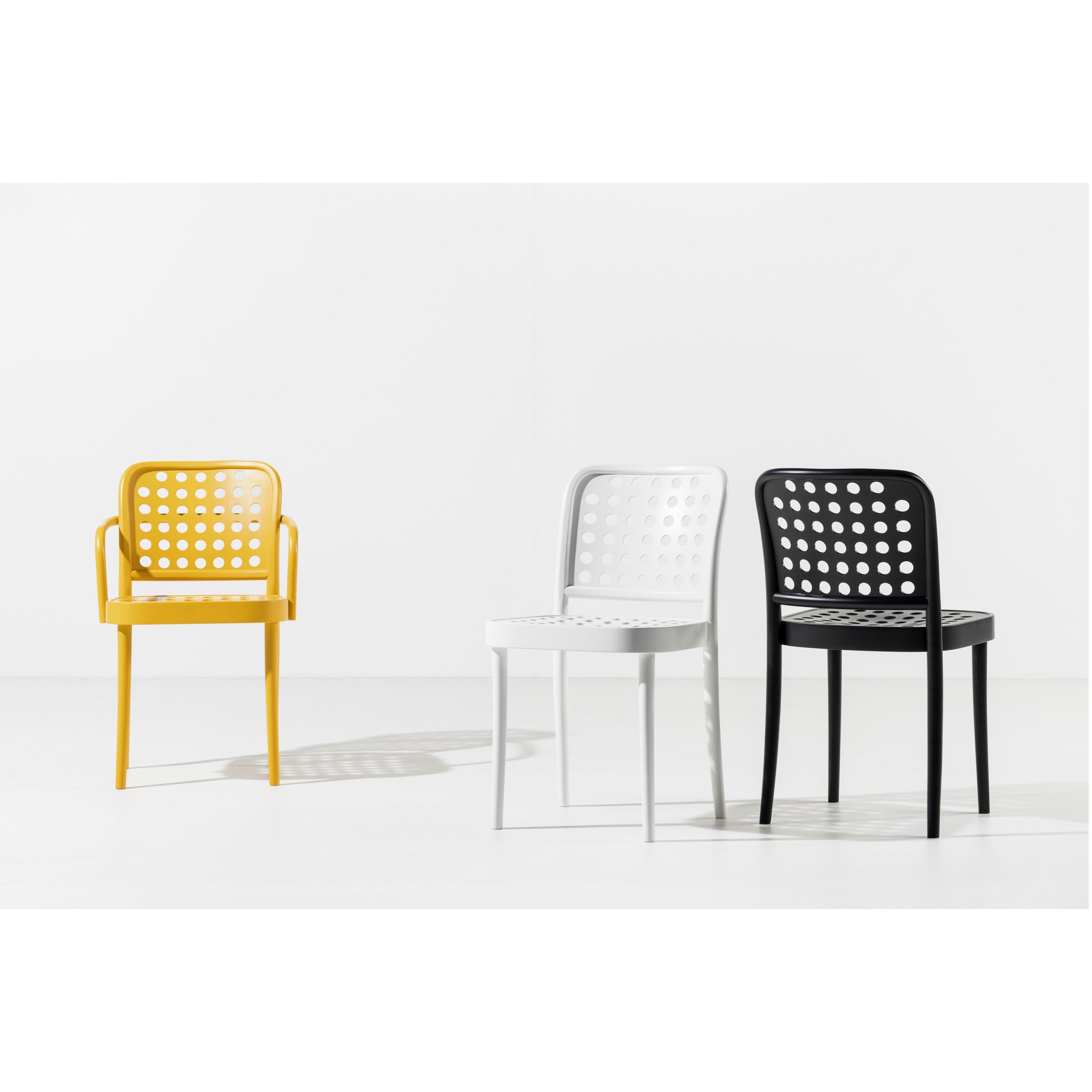 Three perforated TON Chair 822s, epitomizing Scandinavian minimalism, are set against a plain white background. The yellow chair has armrests, while the sleek white and black chairs embrace simplicity without them.