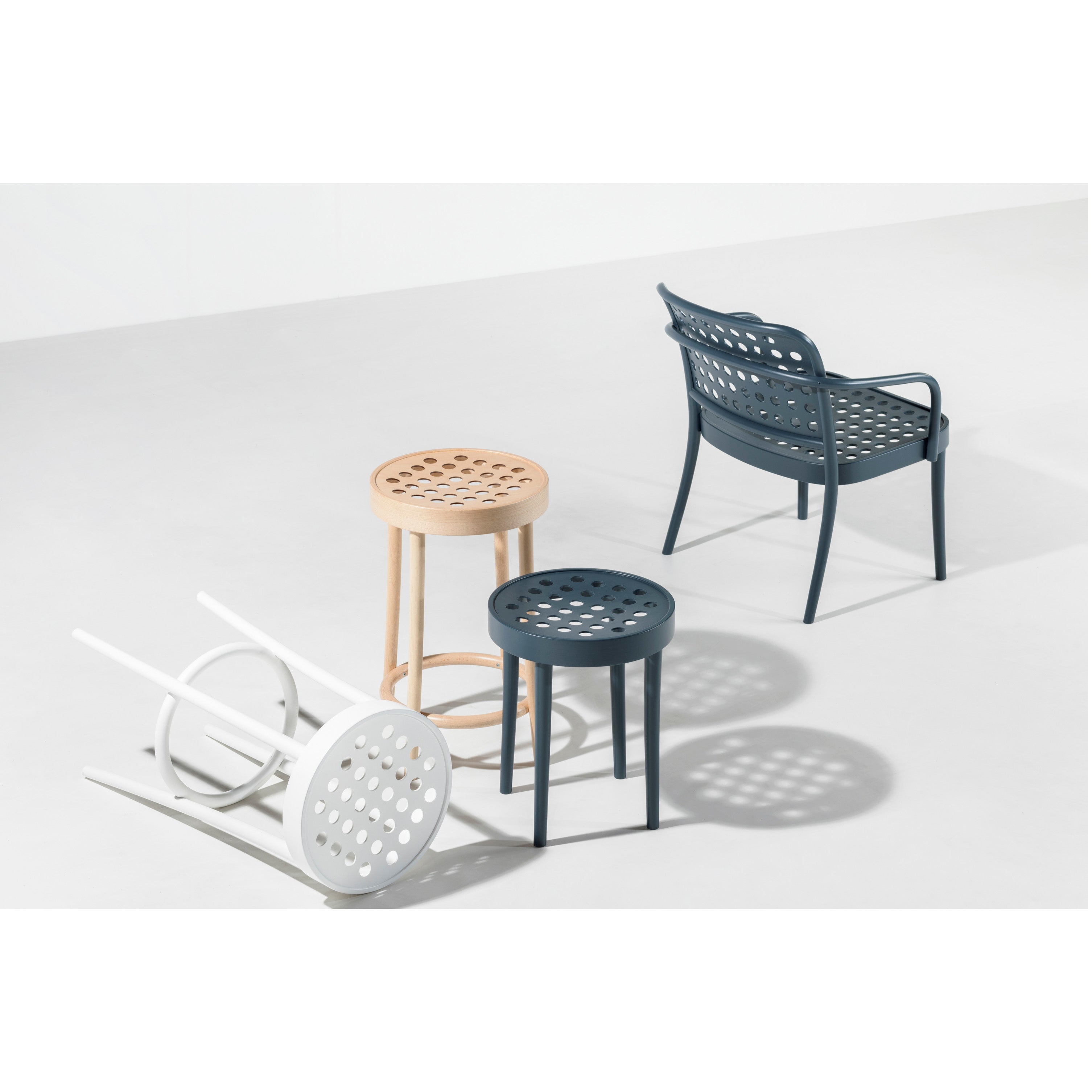 Four stools and a dark TON Armchair 822 with perforated armrests by Claesson Koivisto Rune are arranged on a light surface. Two stools—one wooden, one white—stand upright; another white stool is toppled.