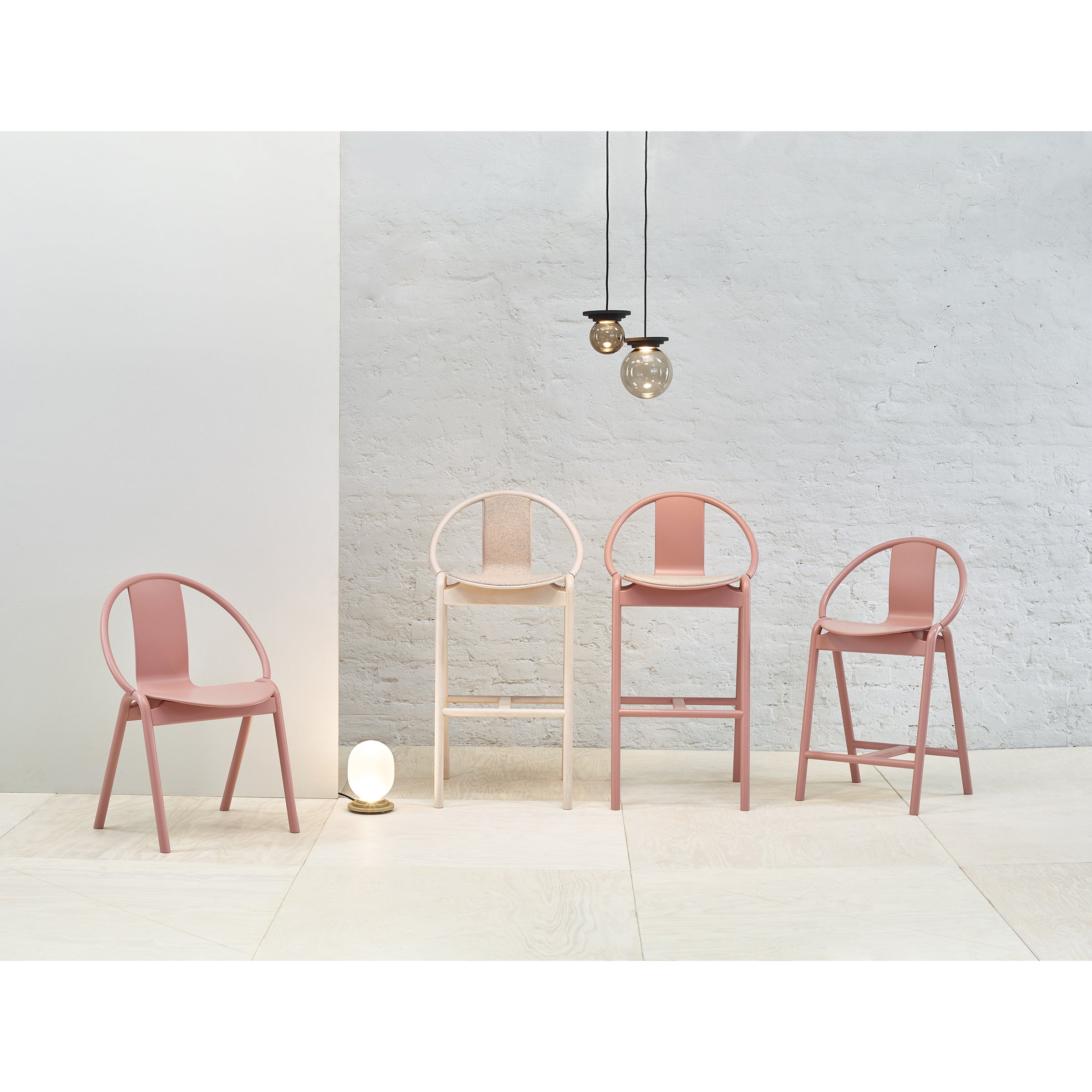 Four modern chairs in varying shades of pink and beige sit before a white brick wall. Overhead, three pendant lights emit a soft glow. A TON Barstool Again Upholstered barstool adds an artful touch to the minimalist ambiance, enhanced by a round floor lamp.