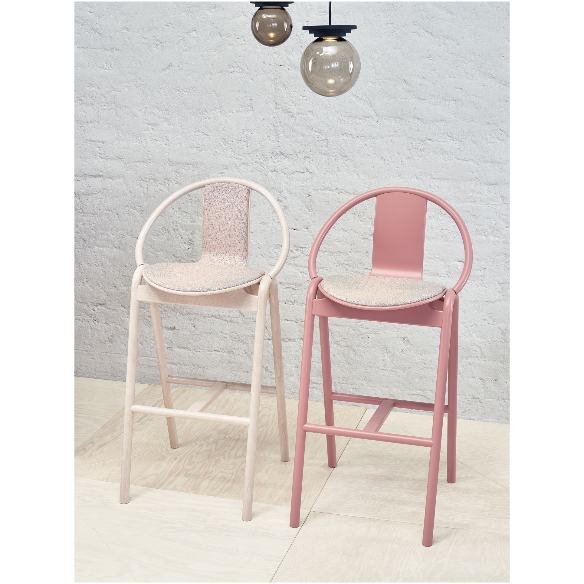 Two Barstool Again Upholstered pieces by TON in muted pink and beige stand on a light wood floor. Overhead, three round pendant lights with black cords complement the textured white brick wall.