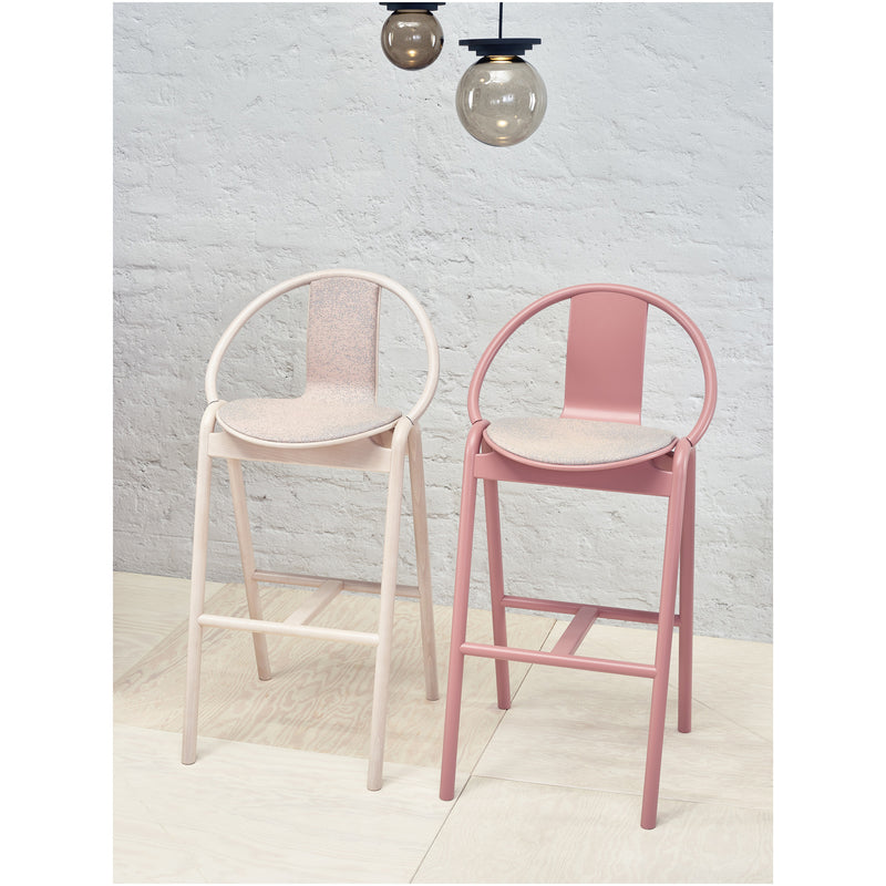 Two Barstool Again Upholstered pieces by TON in muted pink and beige stand on a light wood floor. Overhead, three round pendant lights with black cords complement the textured white brick wall.