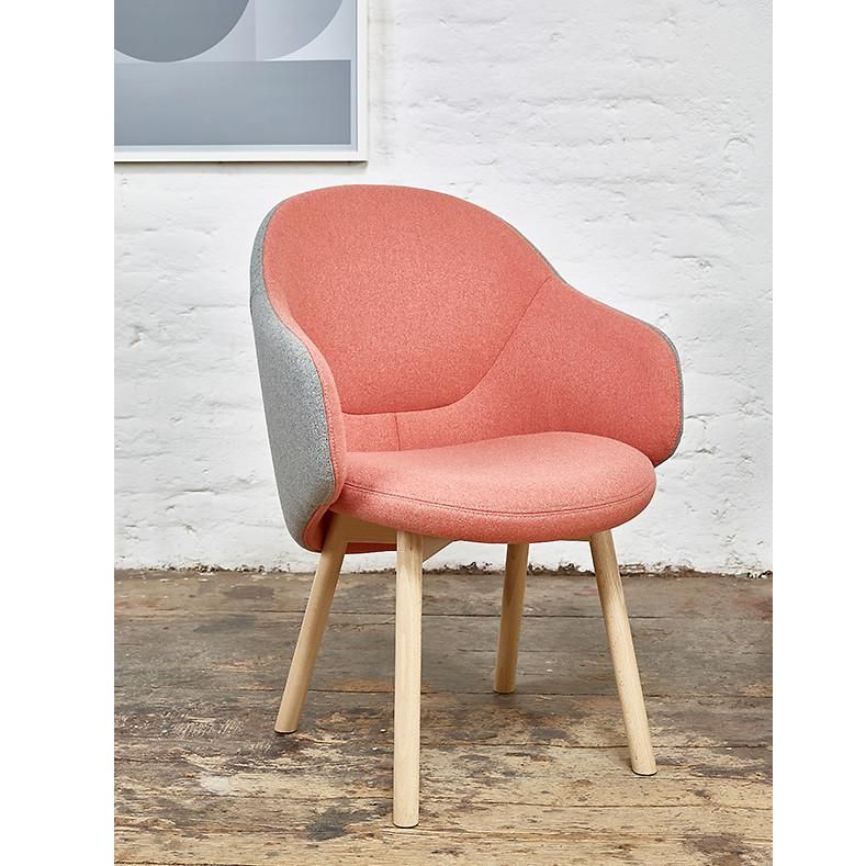 The Armchair Albu by TON, now on sale, showcases an ergonomic design with a pink fabric cushion, gray sides, and light wooden legs. Situated on a wooden floor against a white brick wall, it complements the space where part of a framed abstract print is visible.