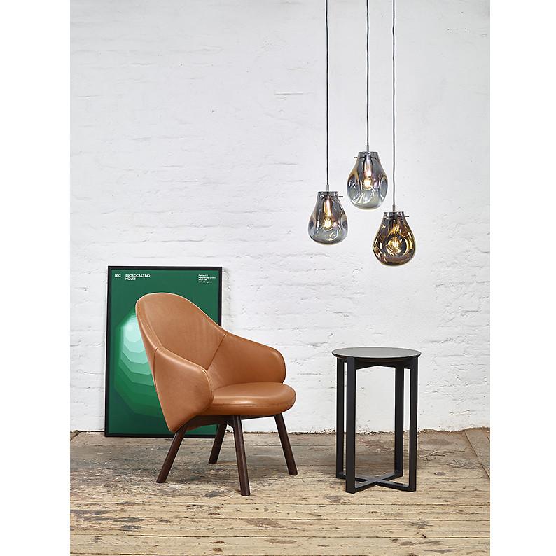 A TON Lounge Armchair Albu in brown pairs with a small black table on a wooden floor. Overhead, three pendant lights add an impressive design element, while a green abstract art piece leans against the white brick wall in the background.