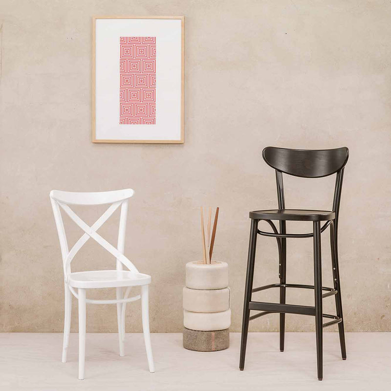 A white chair with an upholstered seat and a black TON Barstool Banana stands on a light wooden floor against a beige wall. Above hangs geometric red-patterned framed artwork. Two cylindrical beige cushions with reed diffusers are between the chairs, enhancing tranquility.