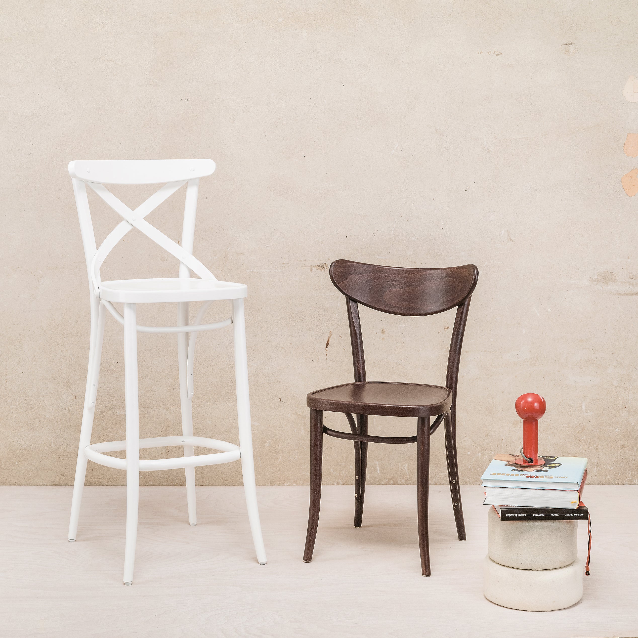 A white bar stool and a TON Chair Banana with an upholstered seat stand on a light wooden floor against a beige wall. A red rubber stamp rests on books atop a round ceramic stool.