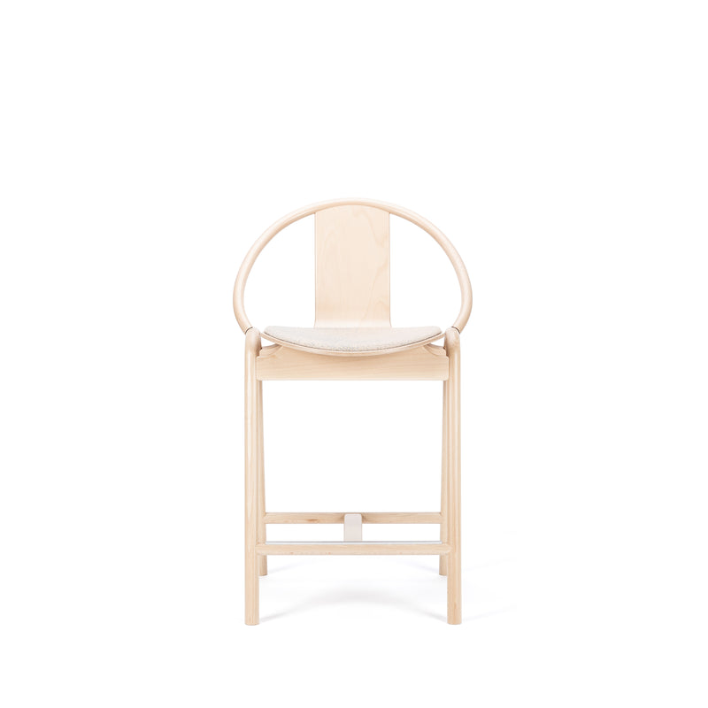 The Barstool Again Upholstered by TON is a minimalist wooden chair with a curved backrest and cushioned seat against a white backdrop. Its light wood finish and sleek, modern design capture the elegance of handcrafted furniture.