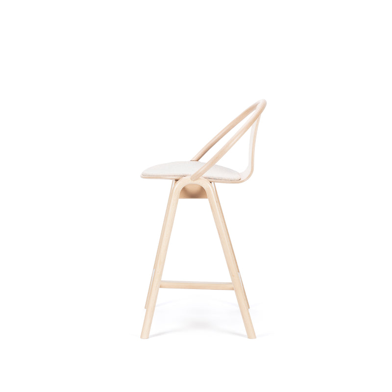The Barstool Again Upholstered by TON is a minimalist wooden chair viewed from the side, featuring a beige cushioned seat, curved backrest, and slender legs. Its handcrafted elegance stands out against the plain white background.