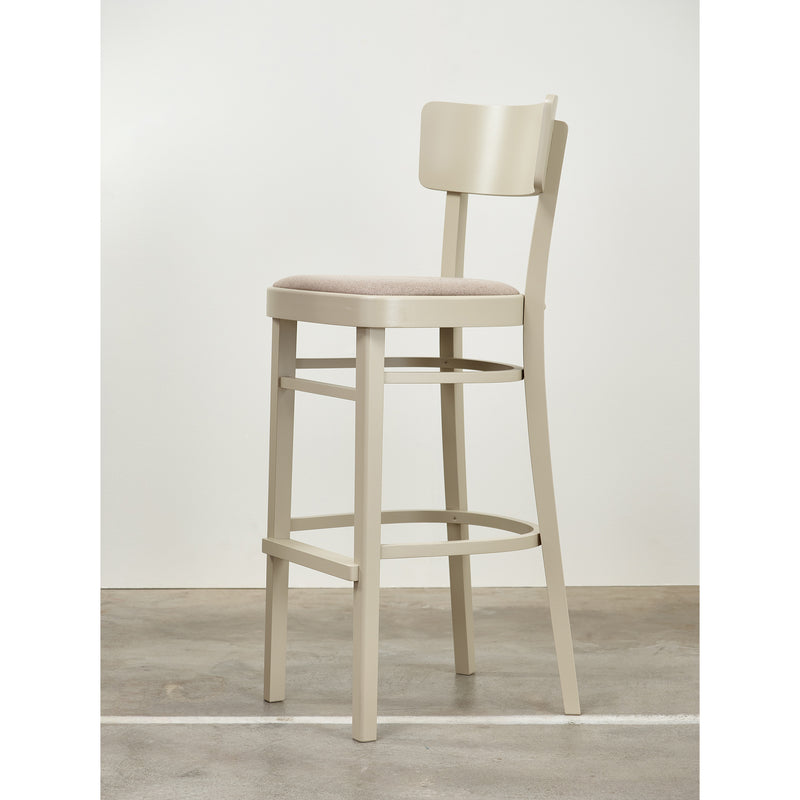 The Barstool Ideal by TON, featuring a curved backrest and cushioned seat, stands on the concrete floor. Its four legs connected by a footrest make it perfect for any modern kitchen or lounge.