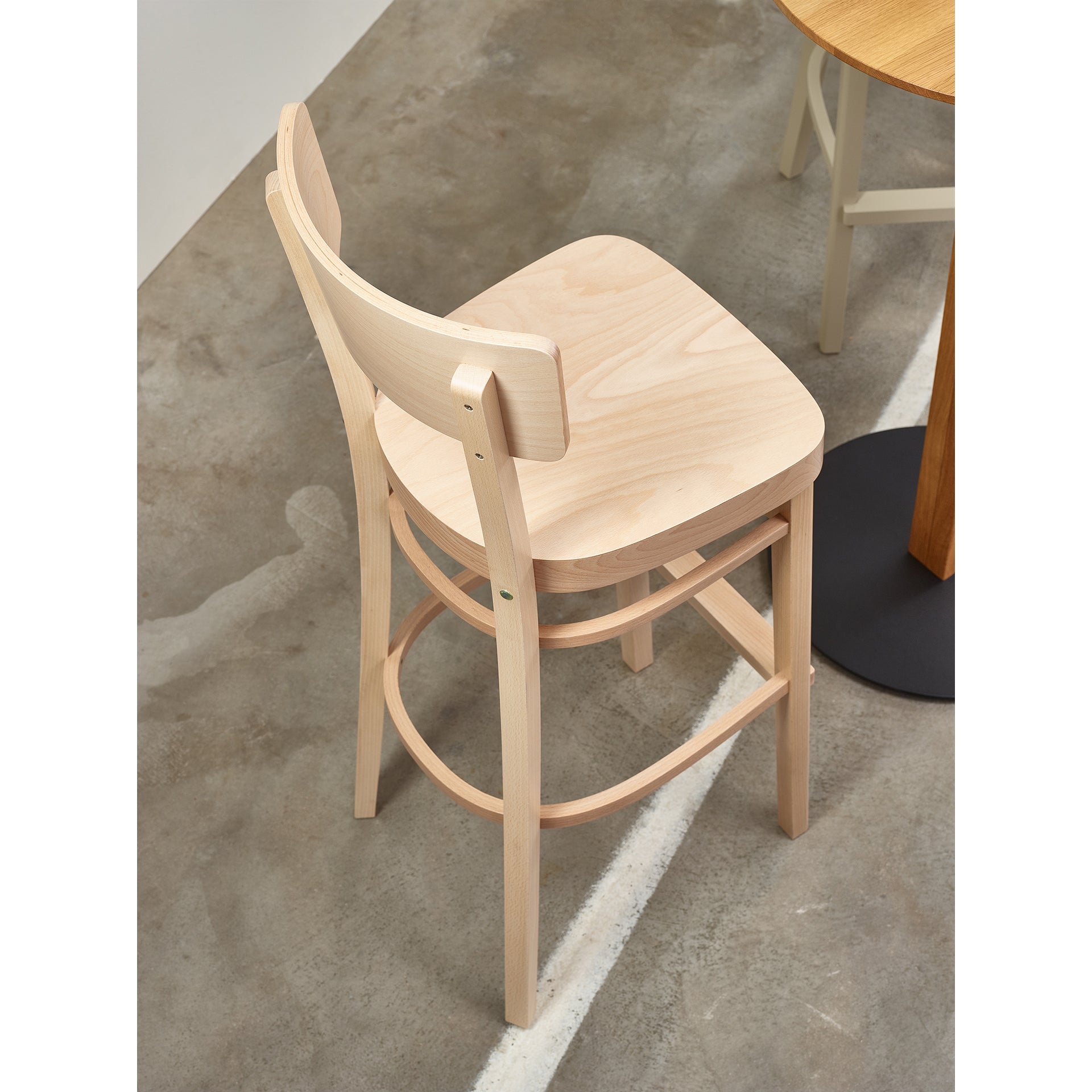 A TON Barstool Ideal with a curved backrest and footrest is placed by a round wooden table on a marked concrete floor, merging style and comfort.