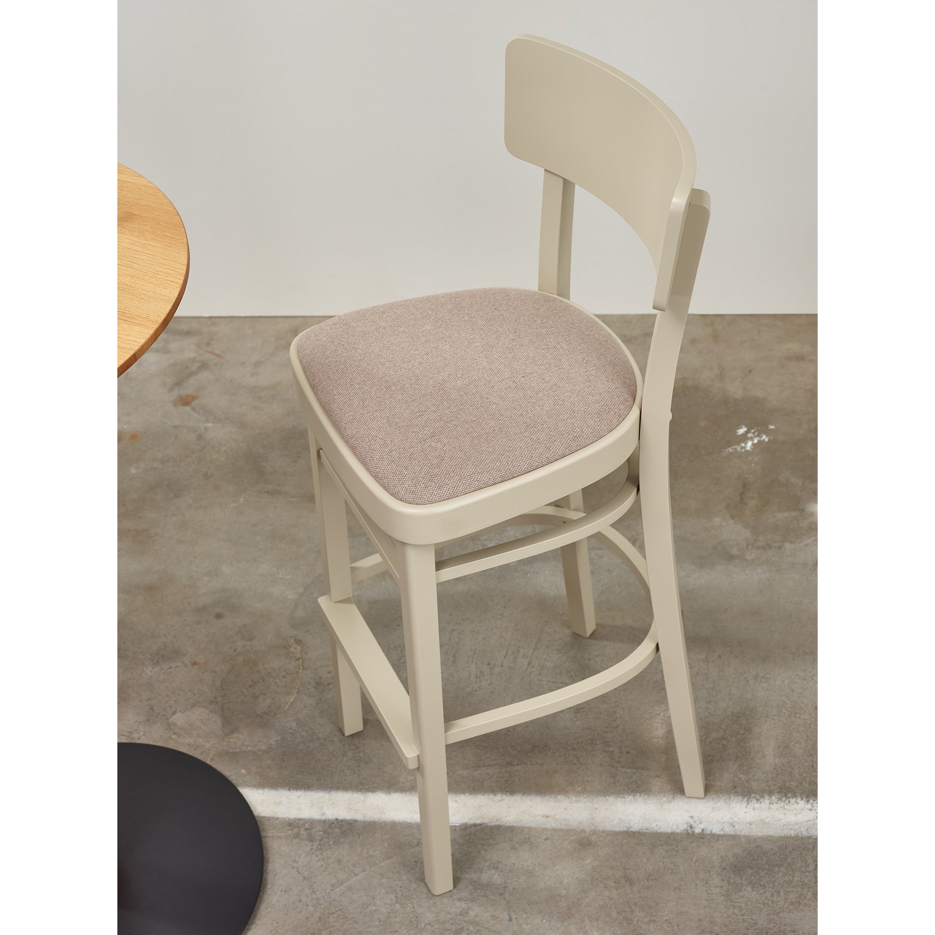 A TON Barstool Ideal with a beige cushion and curved backrest is next to a wooden table. It sits on a concrete floor, with the table edge visible on the left side of the image.