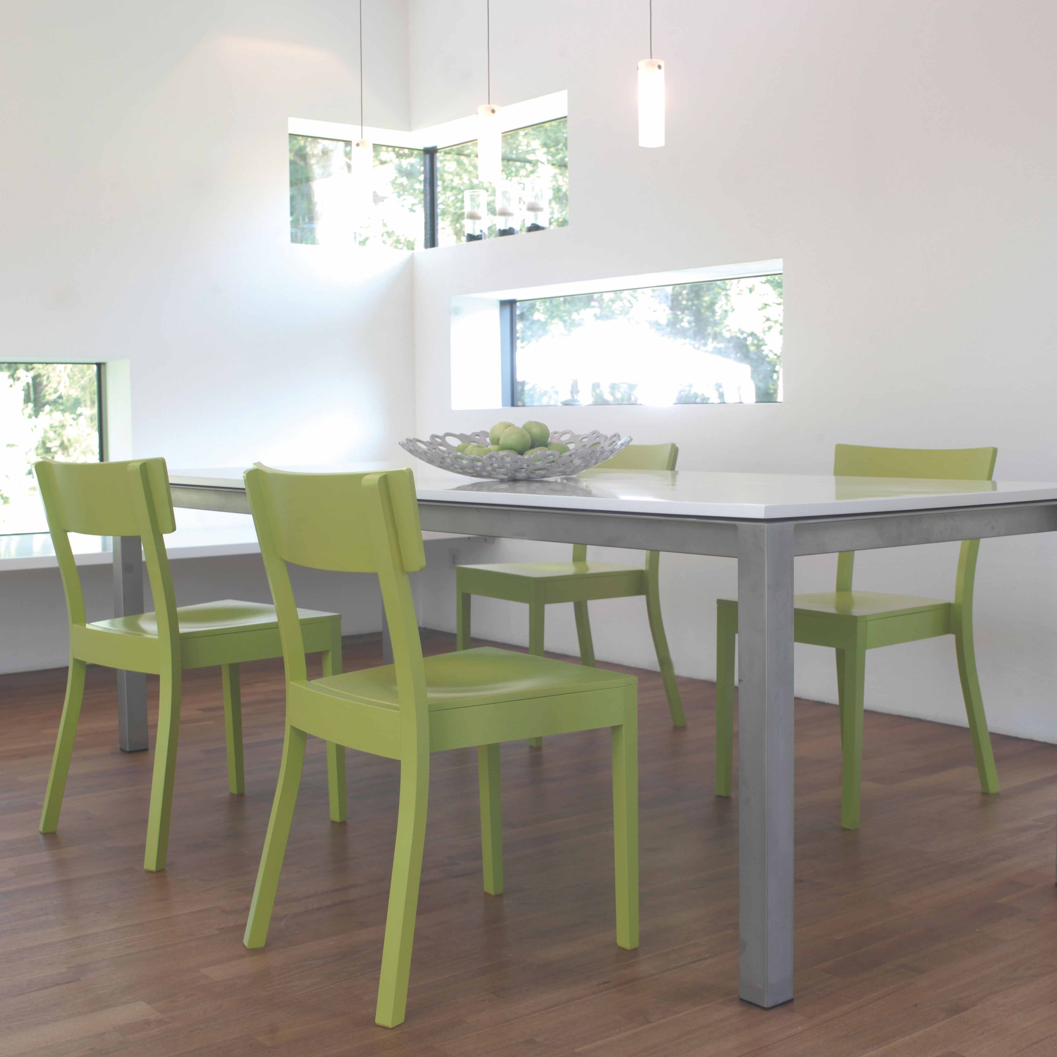 In a modern dining area, the wooden floor showcases a white table and four green TON Chair Bergamo dining chairs. A decorative bowl with green apples embellishes the table. Natural light fills the room through windows in the white walls, with three pendant lights hanging from above.