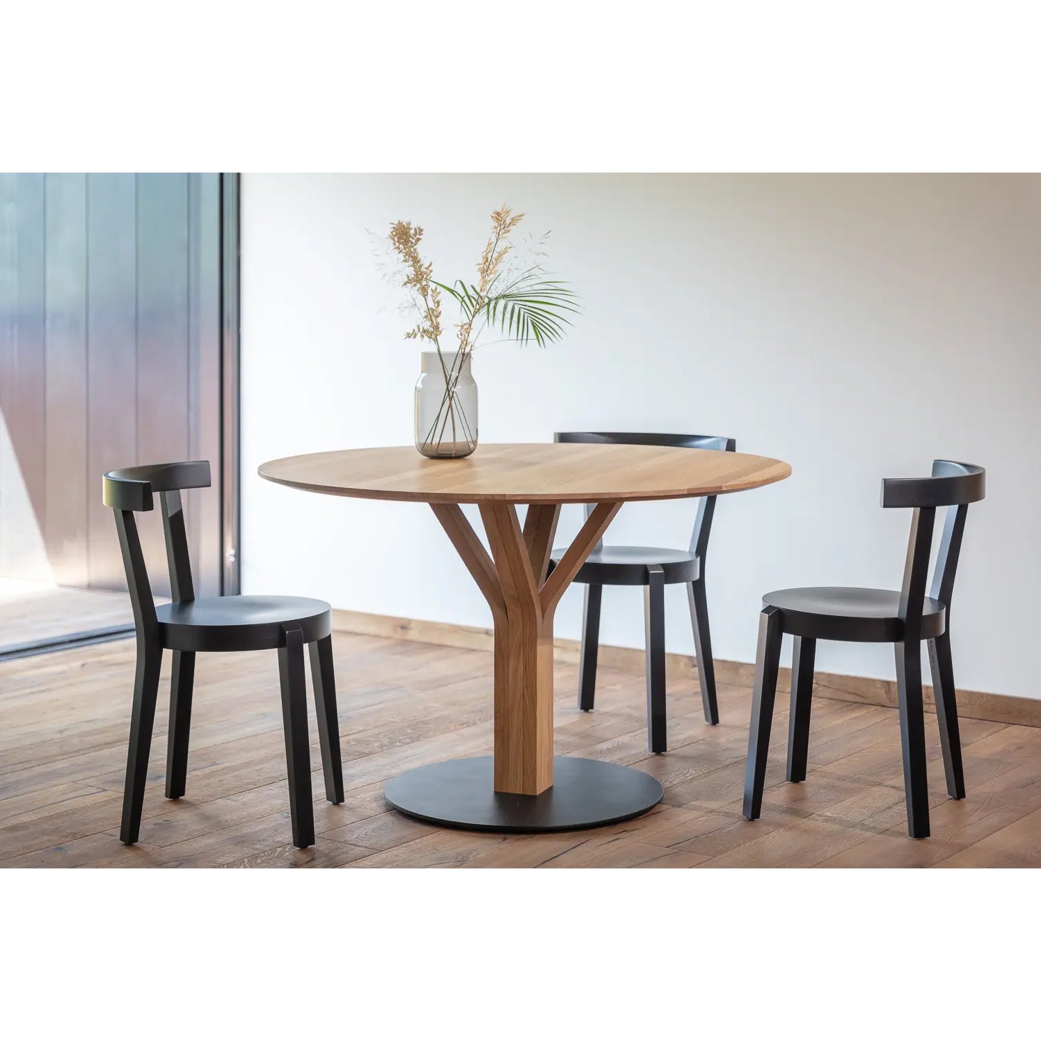 The TON Bloom Central Dining Table, crafted from solid wood with a unique central base, stands on a wooden floor surrounded by three evenly spaced black chairs. A white vase with dried flowers and a palm leaf enhances its elegance as natural light filters through a large window.
