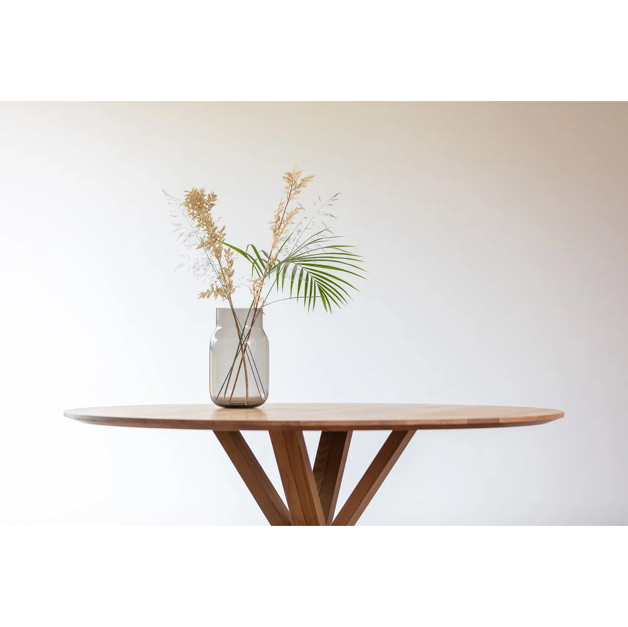 A TON Bloom Central Dining Table with a minimalist design is set against a plain background. Atop it sits a glass vase with dried foliage, such as a palm leaf and wispy stems, accentuating the serene and uncluttered beauty of its solid wood craftsmanship.