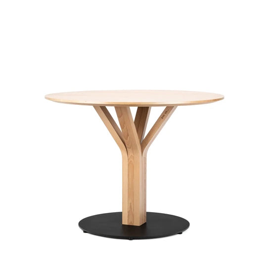 The Bloom Central Dining Table by TON is a round table with a light wood finish, featuring a Y-shaped central support on a solid black circular base. Crafted from solid wood, it stands elegantly against a white background.