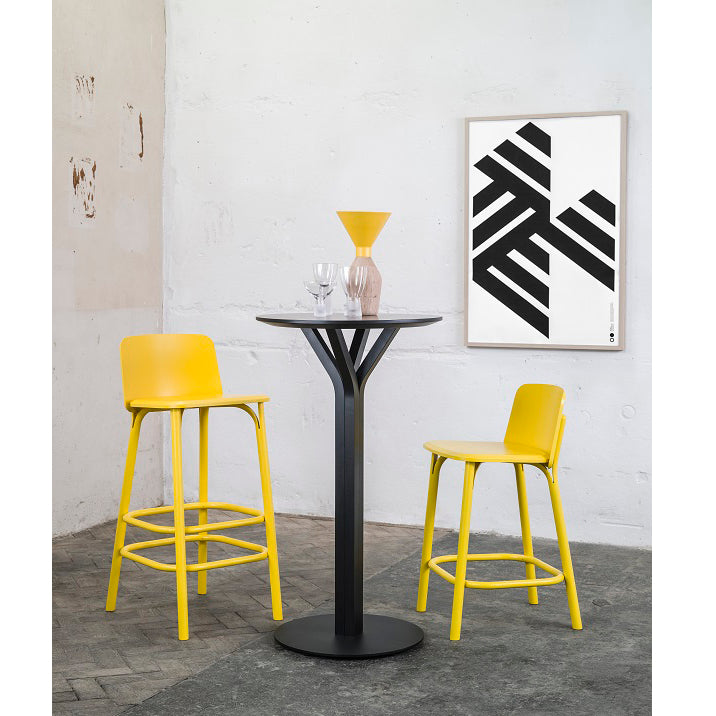 A modern dining setup features the Bloom Bar Table Round by TON, a tall black table with a yellow vase and two glasses. Its paired with two yellow chairs of different heights and highlighted by a black and white geometric art piece on the white wall behind. Crafted from solid wood.