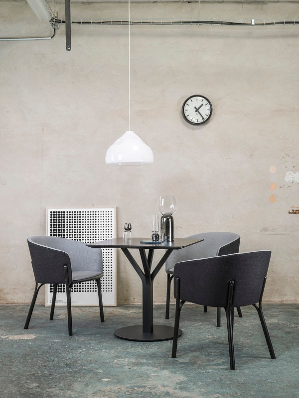 In a minimalist dining area, the Bloom Bistro Table Round by TON is complemented by four modern chairs. Above, a white pendant lamp illuminates the scene. A wall clock discreetly marks time, while textured walls and exposed piping enhance the rooms industrial charm.