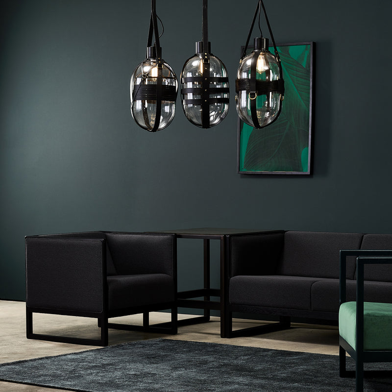 In a modern living room with dark green walls, sleek black sofas and the TON Side Table Casablanca, featuring a veneer or glass top, are showcased. Three hanging glass pendant lights illuminate the green artwork and dark rug, completing the elegant, minimalist design.