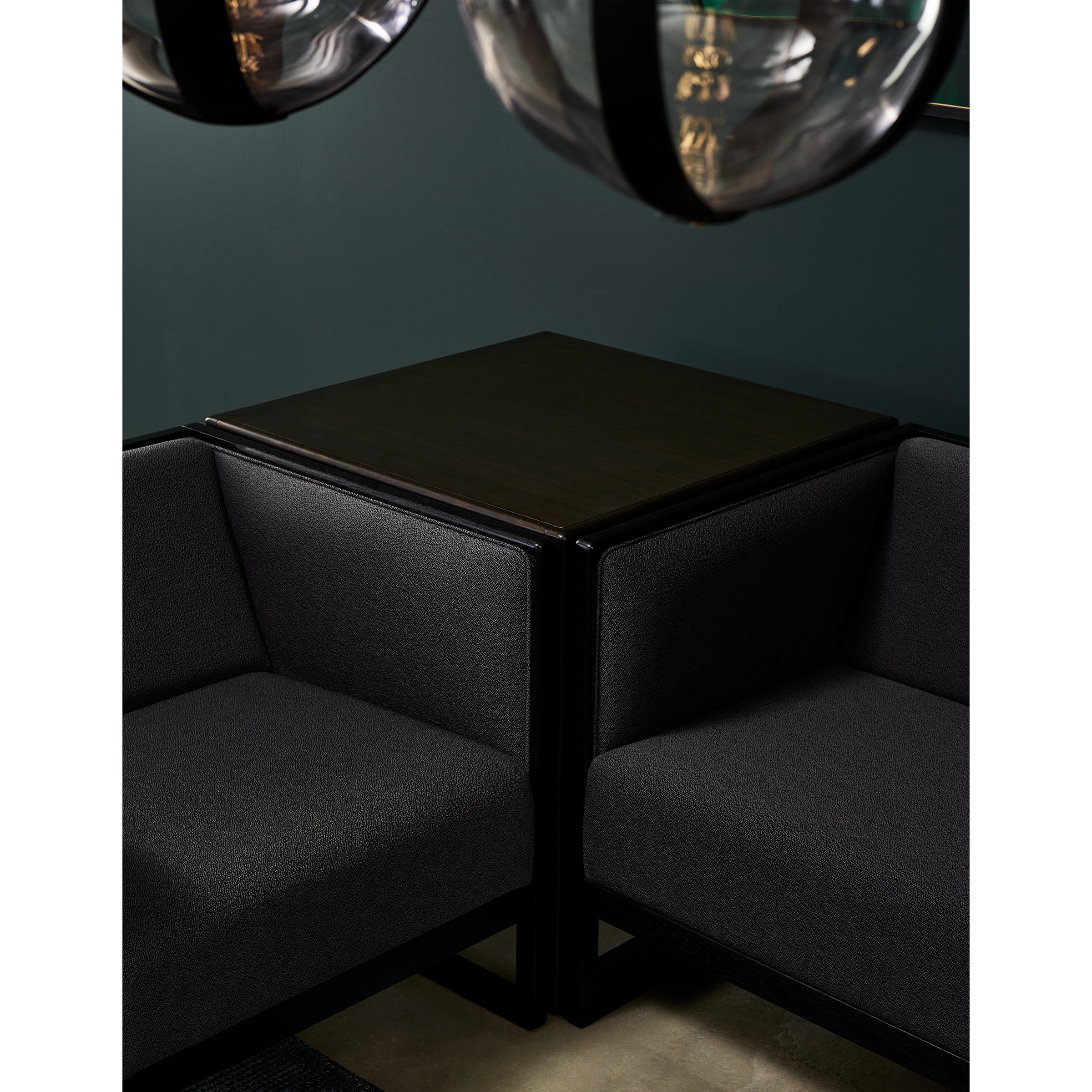 Dark, minimalist seating area with a sleek system of two black couches meeting at the corner around a TON Side Table Casablanca with a glass top. Overhead, two modern, transparent spherical light fixtures with black accents hang against a dark green wall.