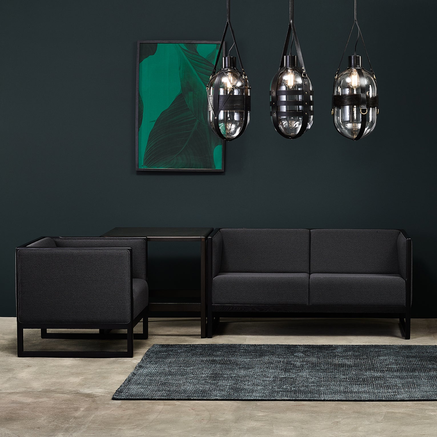 A modern living room showcases dark green walls and a minimalist TON Casablanca Sofa with cane weave armrests. A glass-topped coffee table sits in between, while three industrial-style pendant lights hang above, complemented by a large abstract green artwork on the wall.