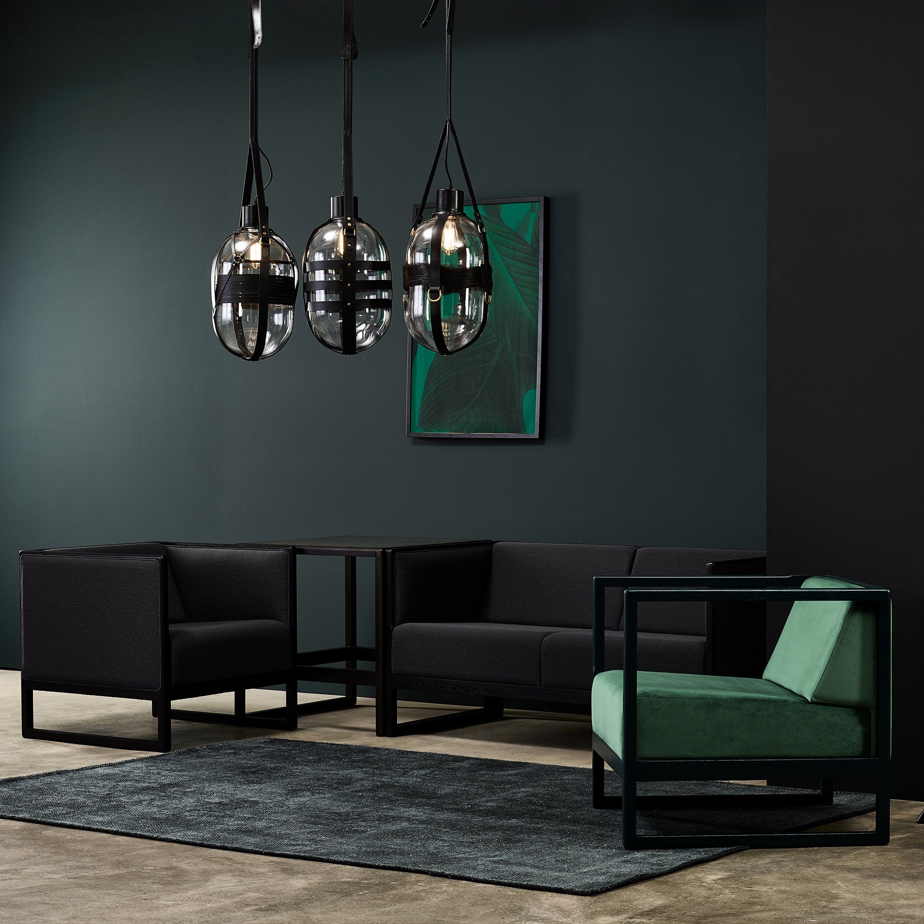A modern living room with dark green walls features three pendant lights, two black armchairs, a TON Casablanca Sofa, a green lounge chair with cane weave armrests, a black table, and a dark rug. A framed abstract artwork gracefully hangs on the wall.