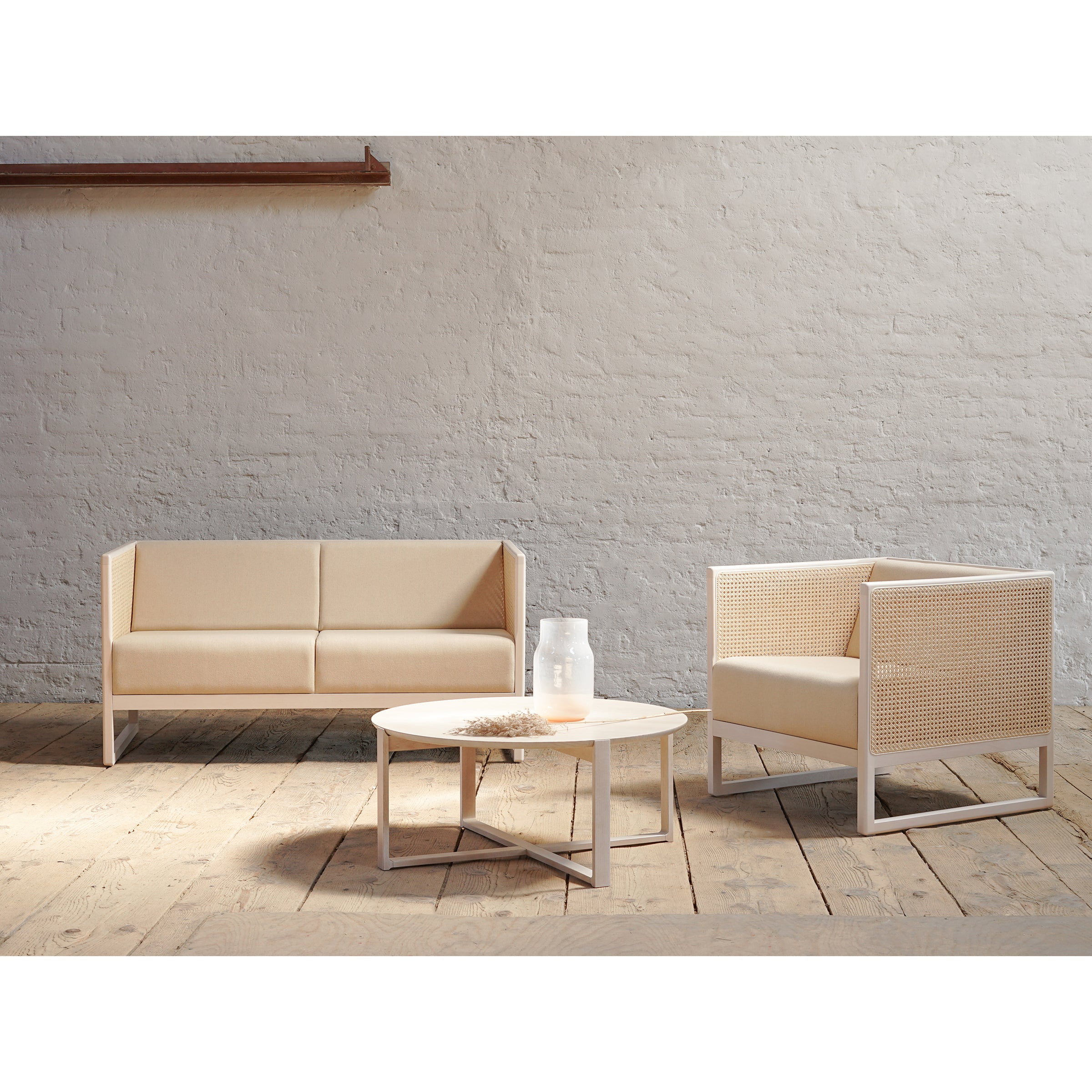 The minimalist living room features a TON Casablanca Sofa and lounge armchair with cane weave armrests against a textured white brick wall. A round coffee table sits in front, holding a glass pitcher and dried florals on the wooden floor.
