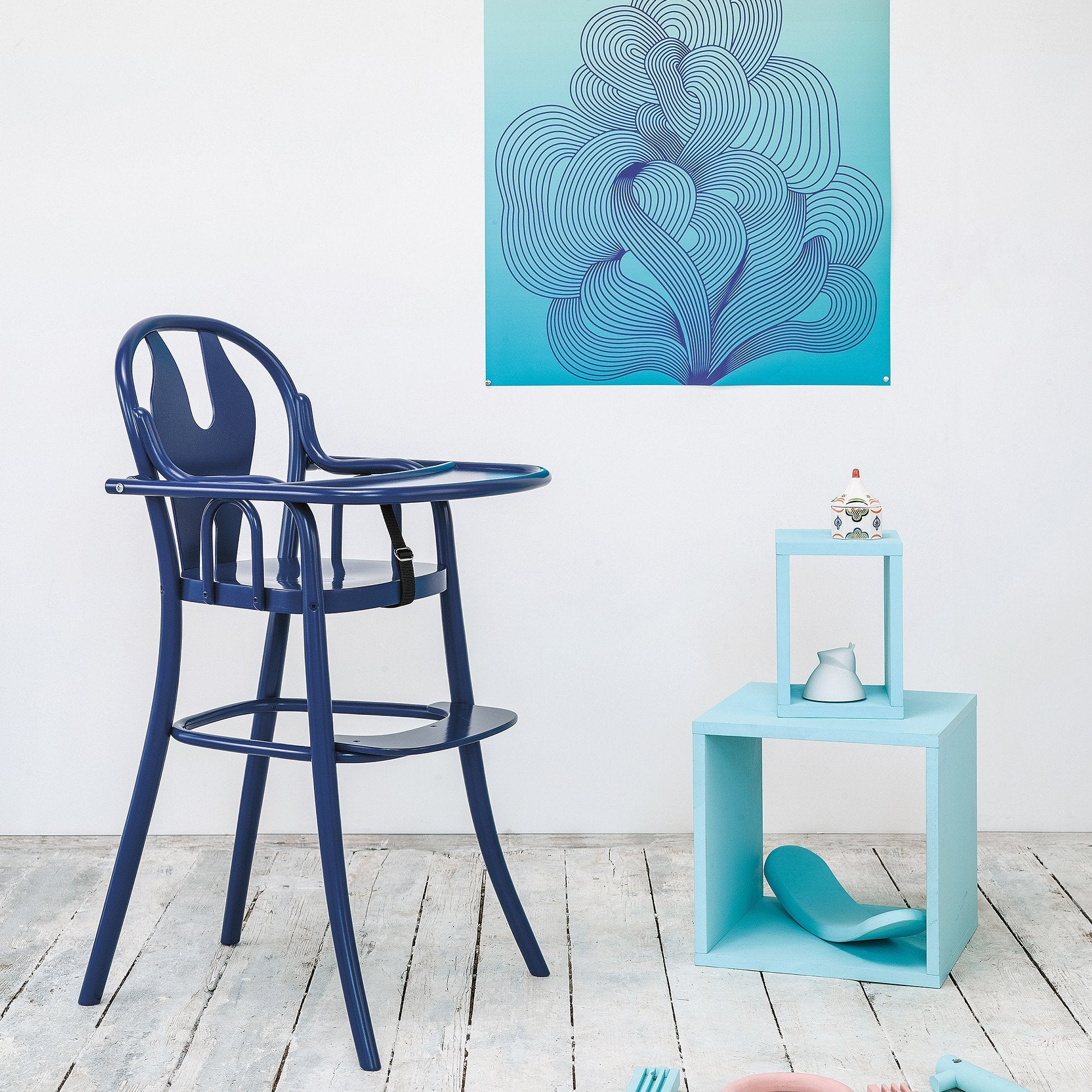 A modern Childrens Chair Petit 114 by TON in dark blue with a safety belt sits on a wooden floor. Nearby, a light blue cube shelf holds ceramic objects. Behind them, a teal abstract swirling artwork adorns the white wall.