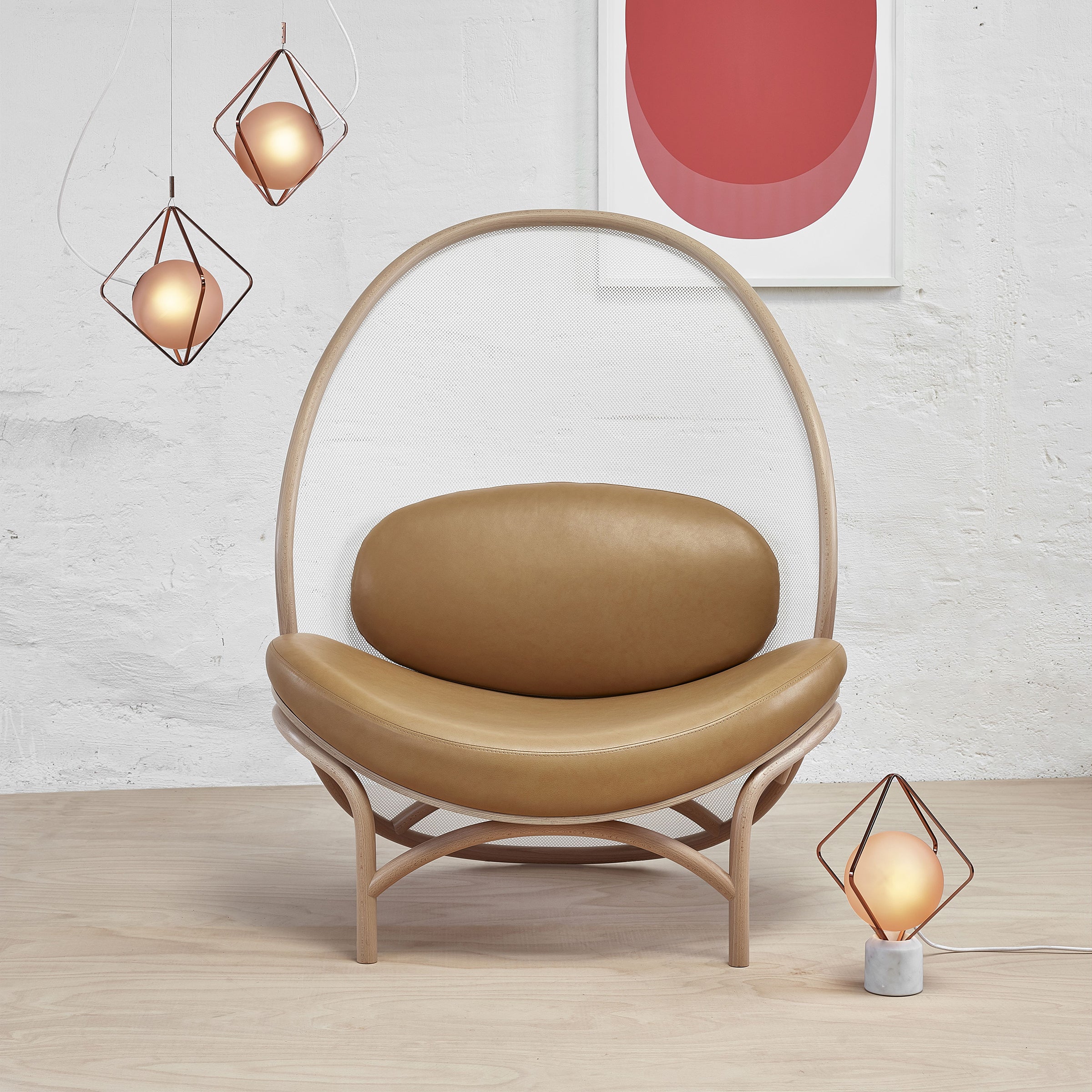The image showcases the Lounge Chair Chips by TON, a stylish modern chair with a curved wooden frame and tan leather upholstery. Nearby are three geometric pendant lights and a matching table lamp with copper accents, while a red and white abstract artwork adorns the wall.