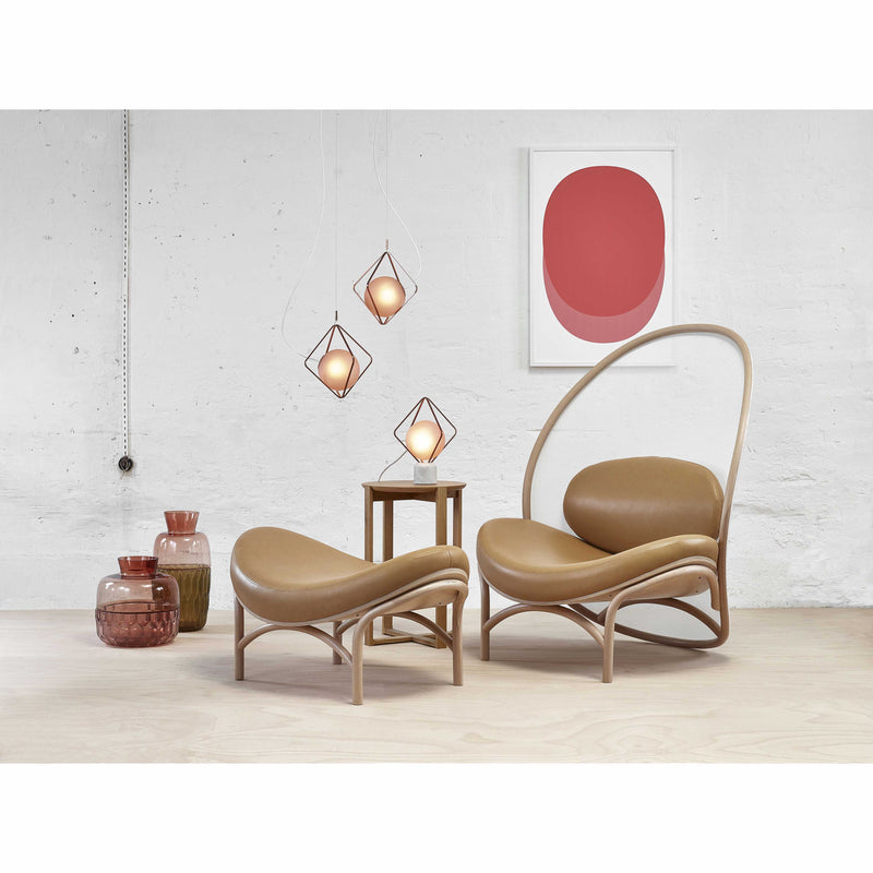 A modern living room showcases a Lounge Chair Chips by TON in tan, featuring elegant manually-bent wood, paired with a matching ottoman on light wood flooring. A small wooden side table holds three pendant lights, red abstract art decorates the white wall, and decorative glass jars sit on the floor.