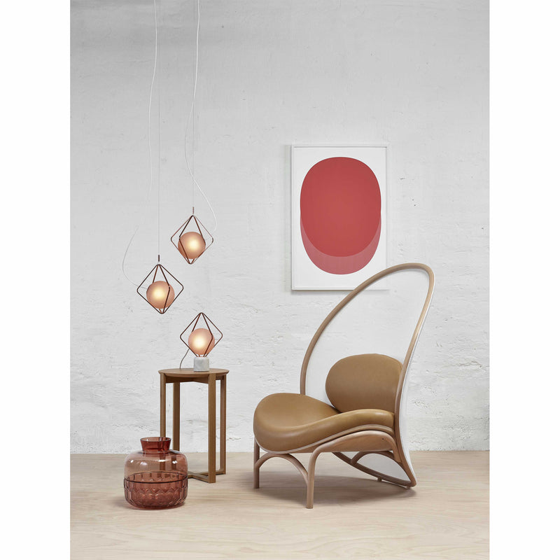 A modern interior features TONs Lounge Chair Chips in tan, boasting cozy upholstery and a curved back. Its paired with a small wooden side table, three geometric pendant lights, and a framed abstract red and pink art piece above a decorative glass jar on the white wall.