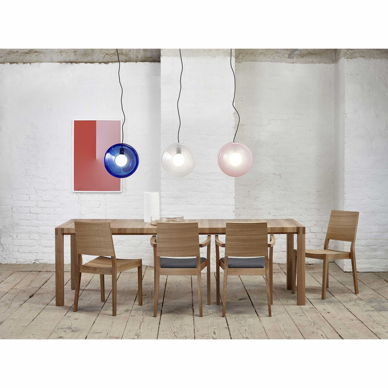 A minimalist dining room showcases a Table Chop by TON, a solid wood table with four chairs on a wooden floor. Above, three pendant lights in blue, clear, and pink hang. A geometric art piece with red and gray squares adorns the white brick wall, exemplifying quality craftsmanship throughout.