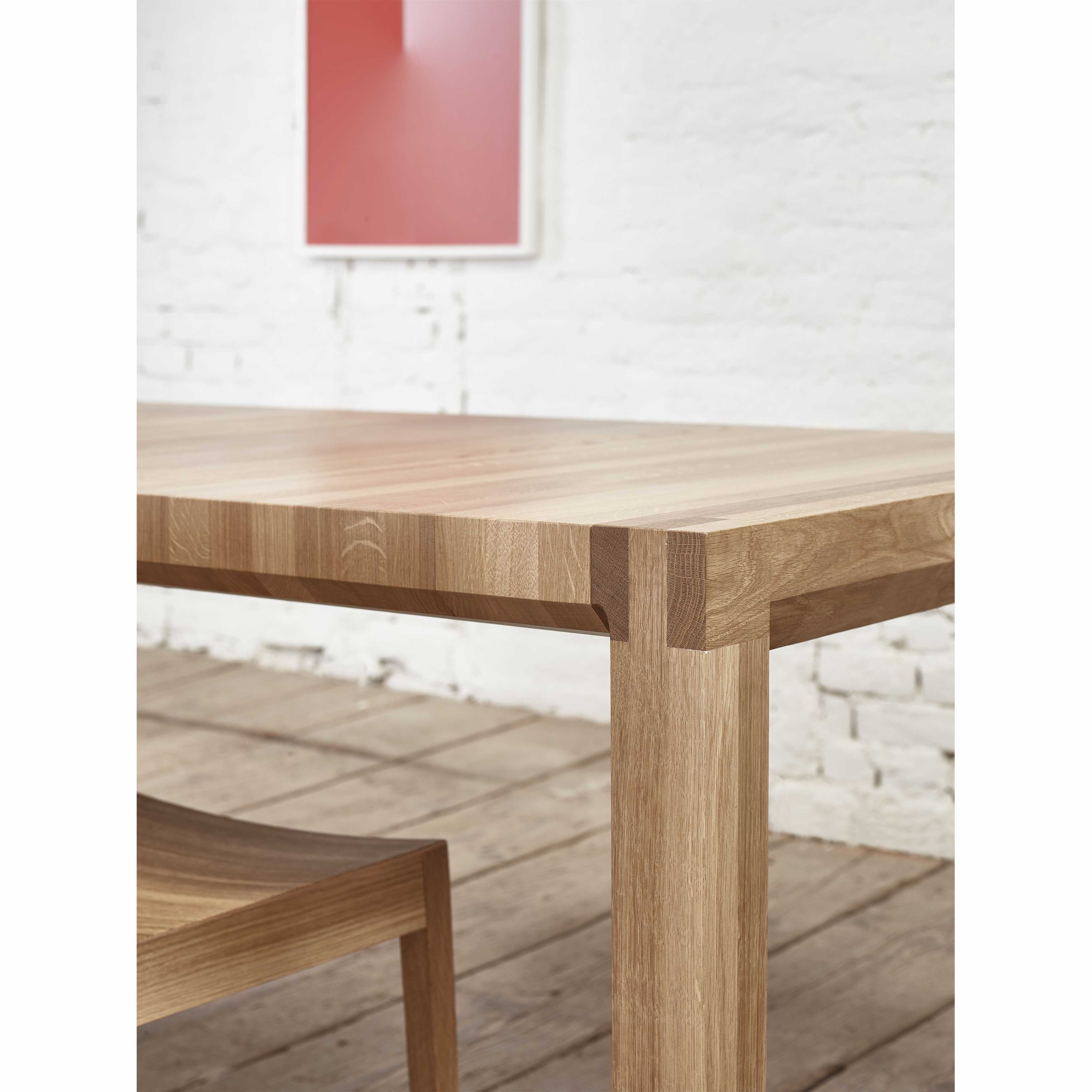 An extendable wooden Table Chop by TON and a matching chair are on a wooden floor, set against a white brick wall with framed abstract red and white art, showcasing minimalist modern quality craftsmanship.