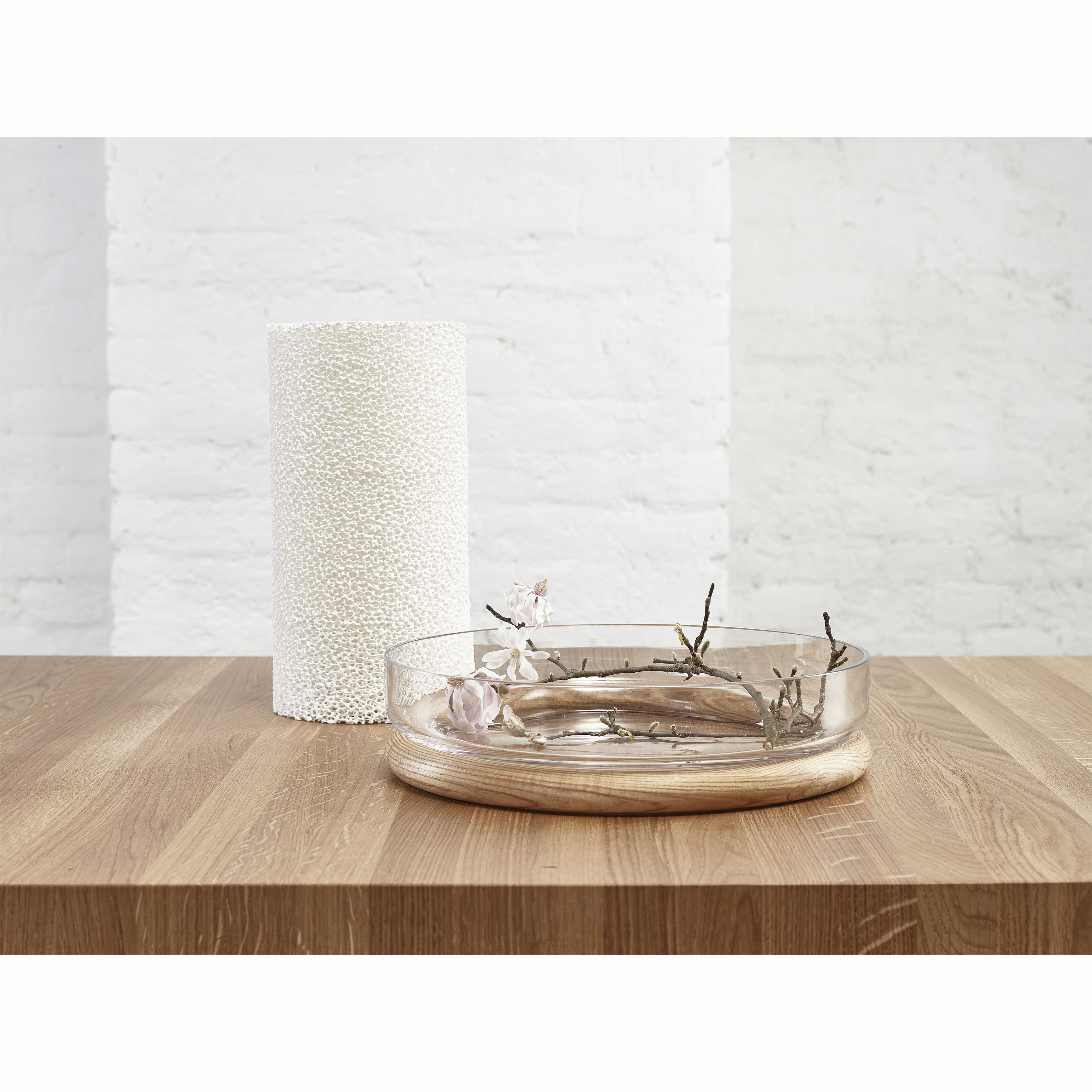 In a minimalist scene, a shallow glass bowl with delicate branches and white flowers sits elegantly on the TON Table Chop, a solid wood table. Beside it, a textured white cylindrical object stands against a white brick wall, representing quality craftsmanship.