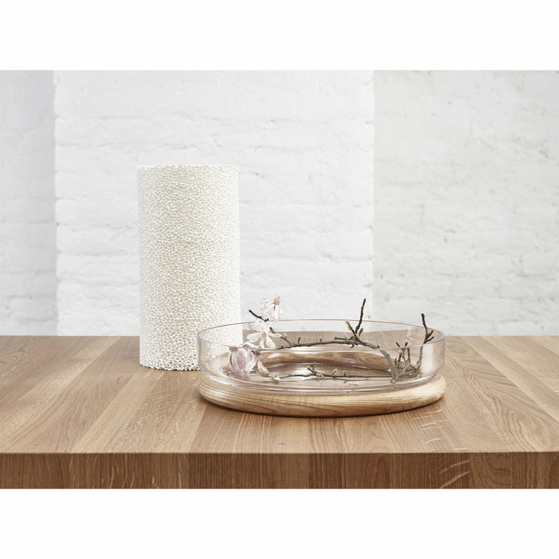 In a minimalist scene, a shallow glass bowl with delicate branches and white flowers sits elegantly on the TON Table Chop, a solid wood table. Beside it, a textured white cylindrical object stands against a white brick wall, representing quality craftsmanship.