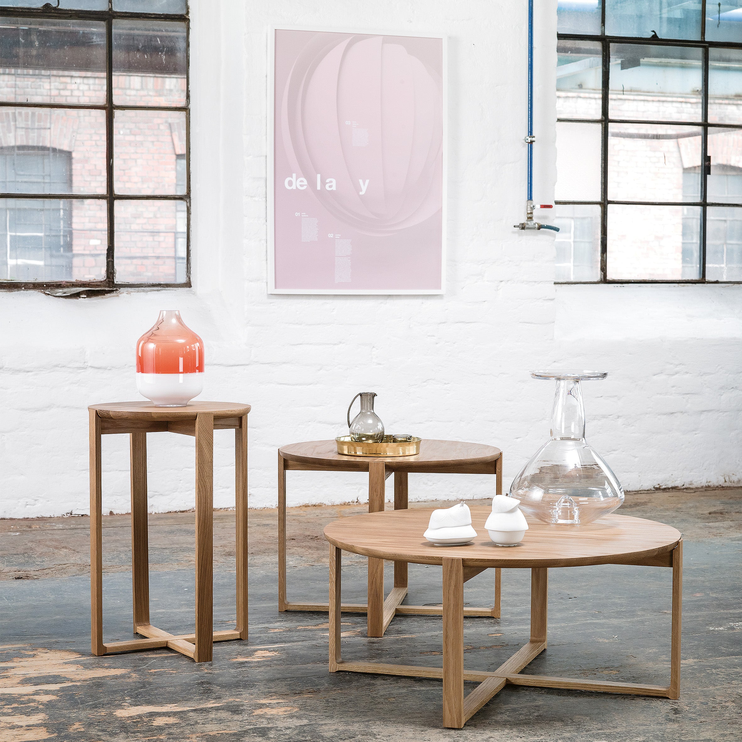 A minimalist room features three Delta Coffee Tables by TON of varying heights. These wooden tables display decorative glass and ceramic items. A framed art piece adorns the white brick wall, while large industrial-style windows flood the space with natural light.