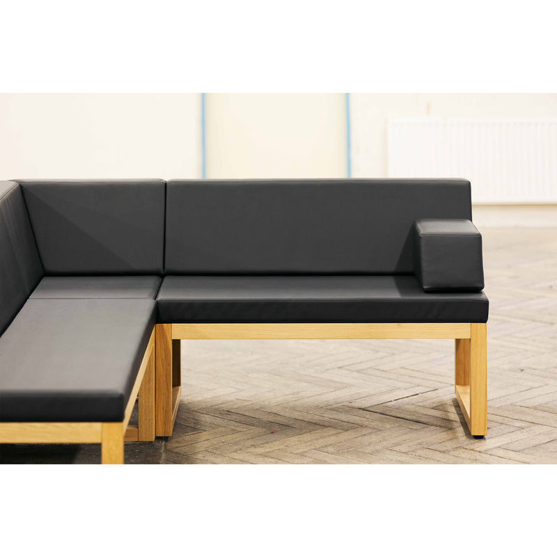The Bench Diner, designed by Kai Stania for TON, is a black leather L-shaped sectional with wooden framing. It sits on a herringbone floor and features a small cushioned armrest. A subtle white background with a faint radiator completes the versatile seating appeal.