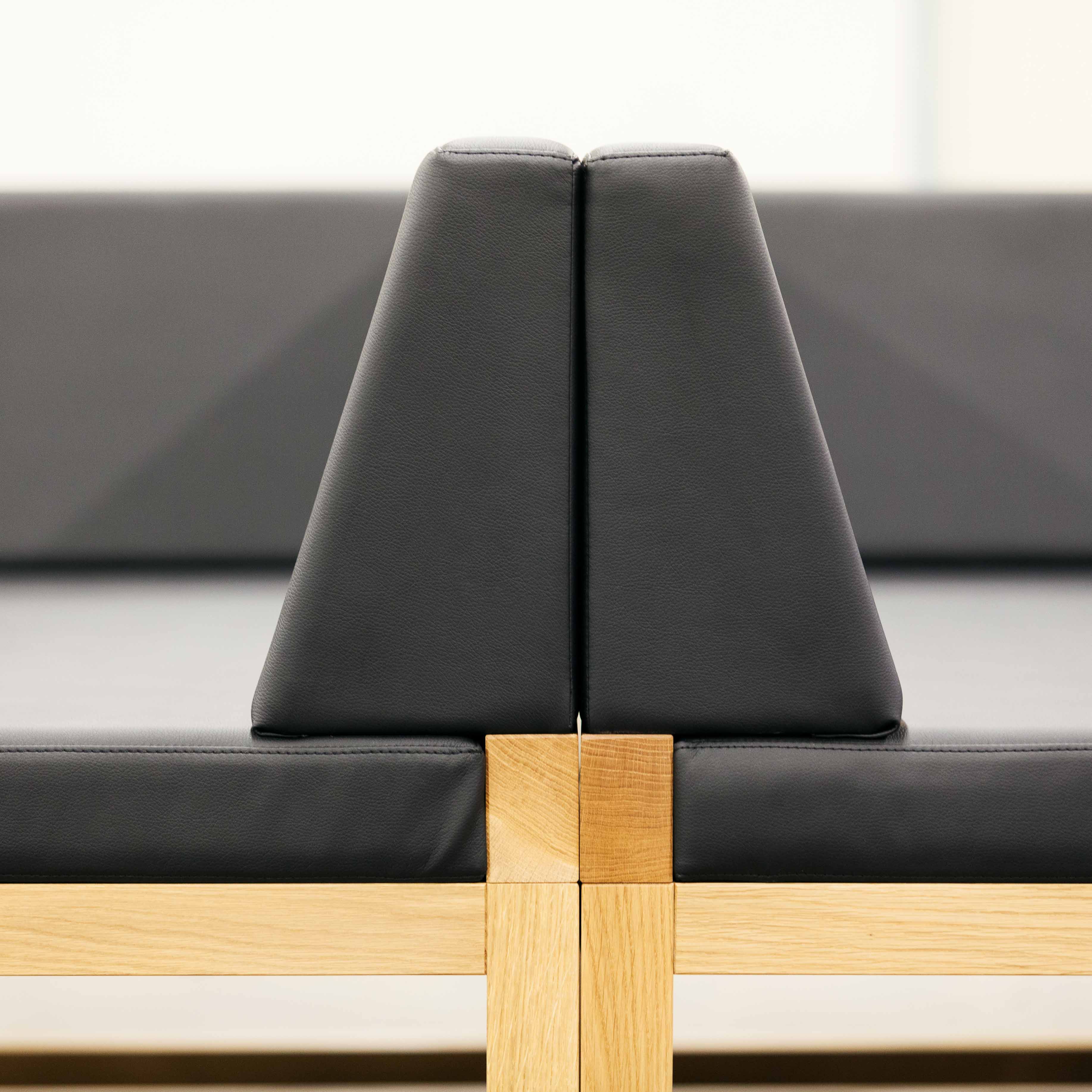 Close-up of the Bench Diner by TON, a modern modular bench system featuring a wooden frame and black cushioned seats. Two triangular backrests converge at the center for a striking geometric design. The softly blurred background highlights its elegance and versatility, crafted by Kai Stania.