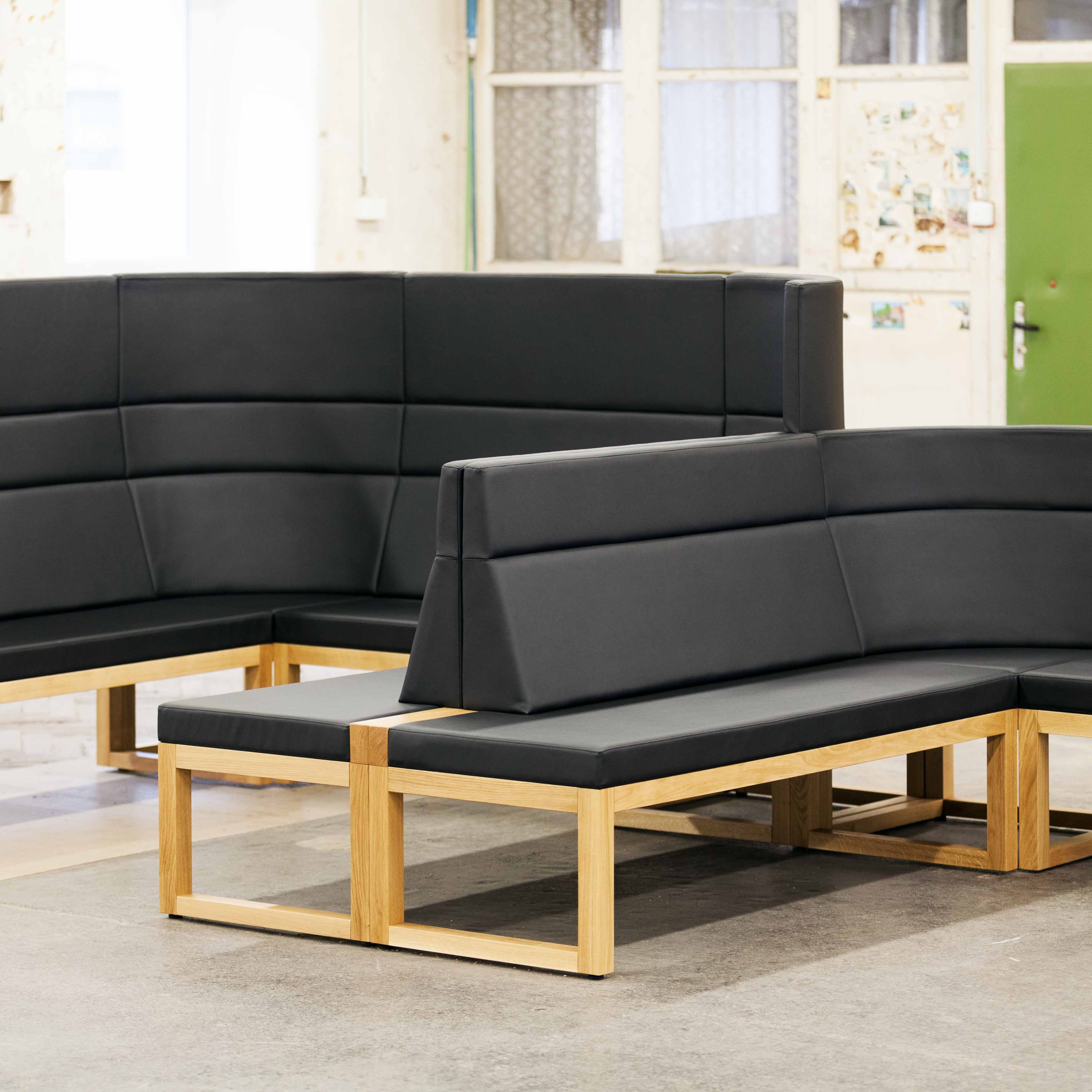 The Bench Diner by TON, inspired by Kai Stania, features black cushions and wooden frames in a bright setting. This versatile seating forms an angular layout against white walls, windows, and a green door.