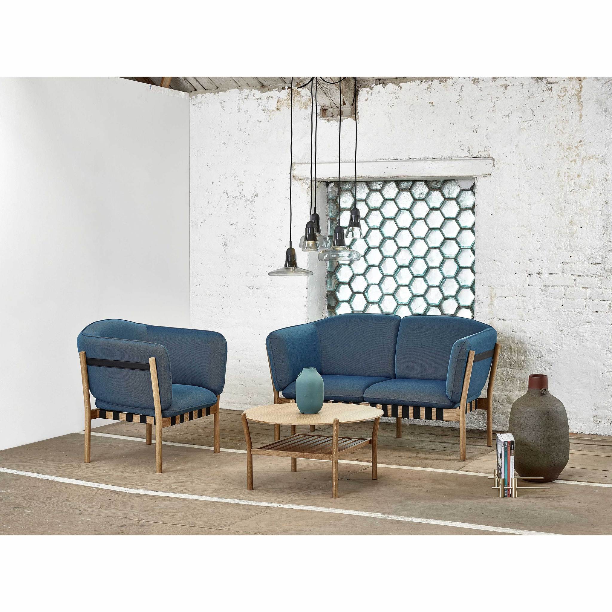 A contemporary room features a comfortable seating area with a TON Dowel Two-Seater and a blue armchair, both with wooden frames. It has white brick walls, hexagonal-patterned windows, hanging pendant lights, and a small wooden coffee table holding a blue vase alongside a large ceramic vase.