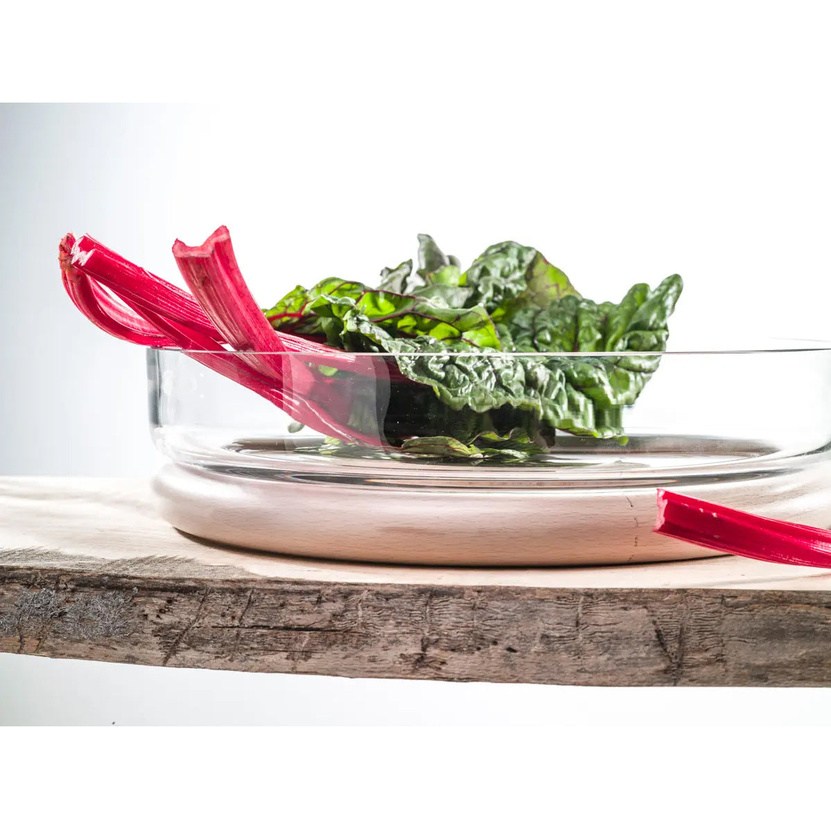 A glass Hoop Bowl by TON, crafted from exquisite Czech crystal glass, holds vibrant green Swiss chard leaves with long red stalks against a rustic wooden surface and soft, light background.