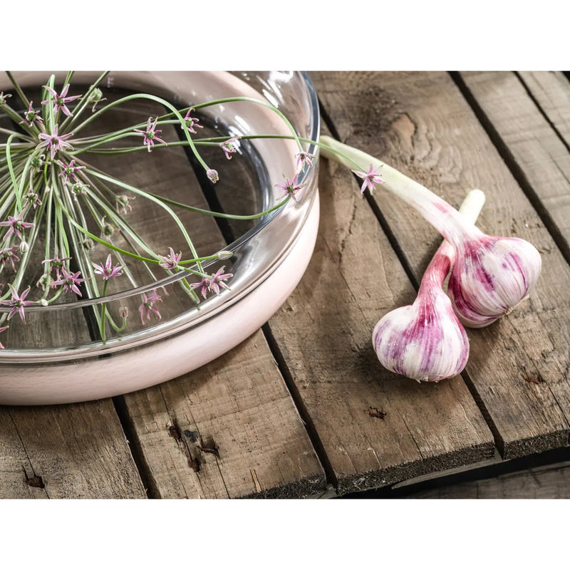 Fresh garlic bulbs with purple streaks rest on a rustic wooden surface beside a TON Hoop Bowl, where delicate pink and white flowers, likely garlic blossoms, elegantly adorn the rim.