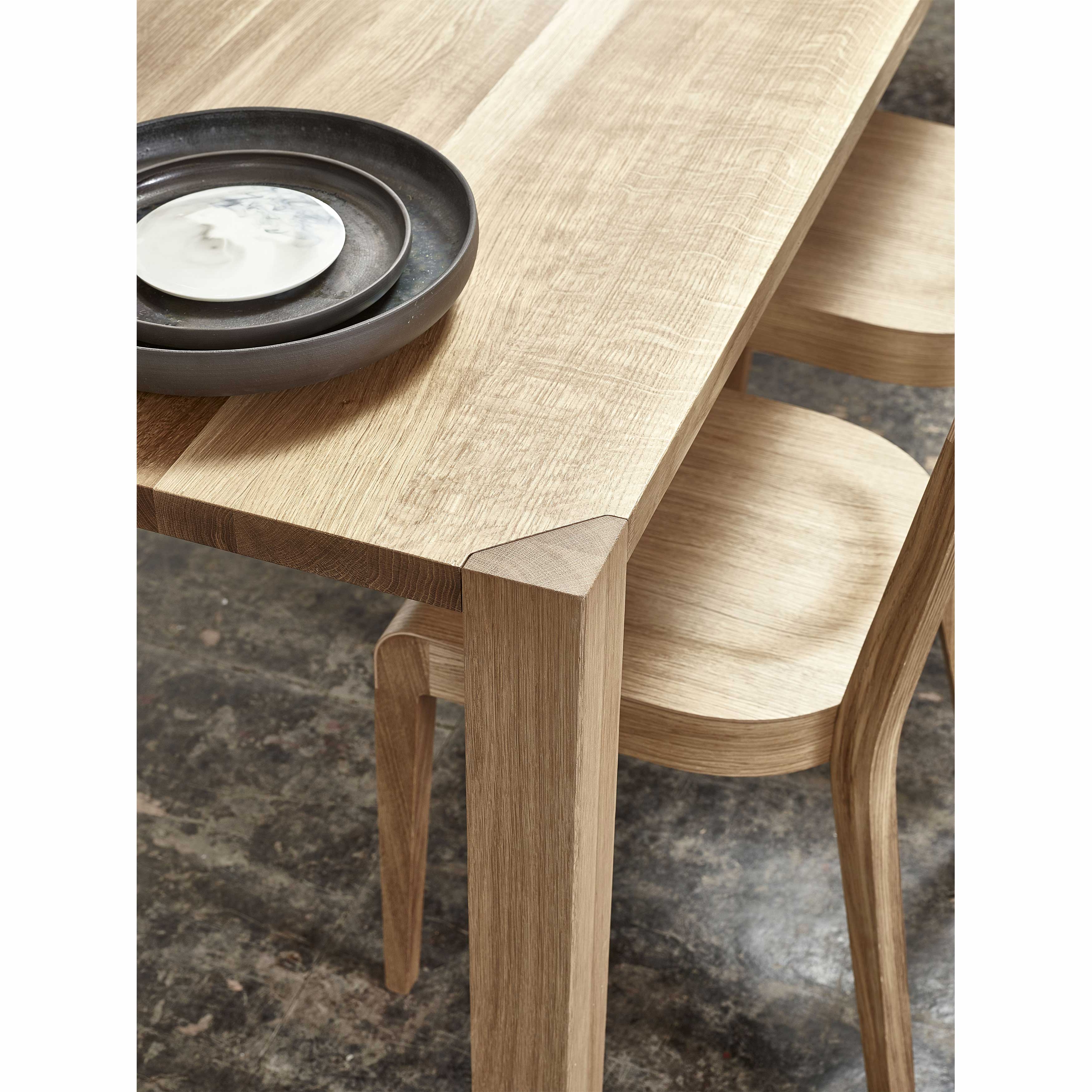 A close-up of the Table Ink dining set by TON shows a thin tabletop with sleek, modern lines on a polished concrete floor. Stacked black ceramic plates and a white saucer highlight the wood grain texture against its elegant triangular legs.