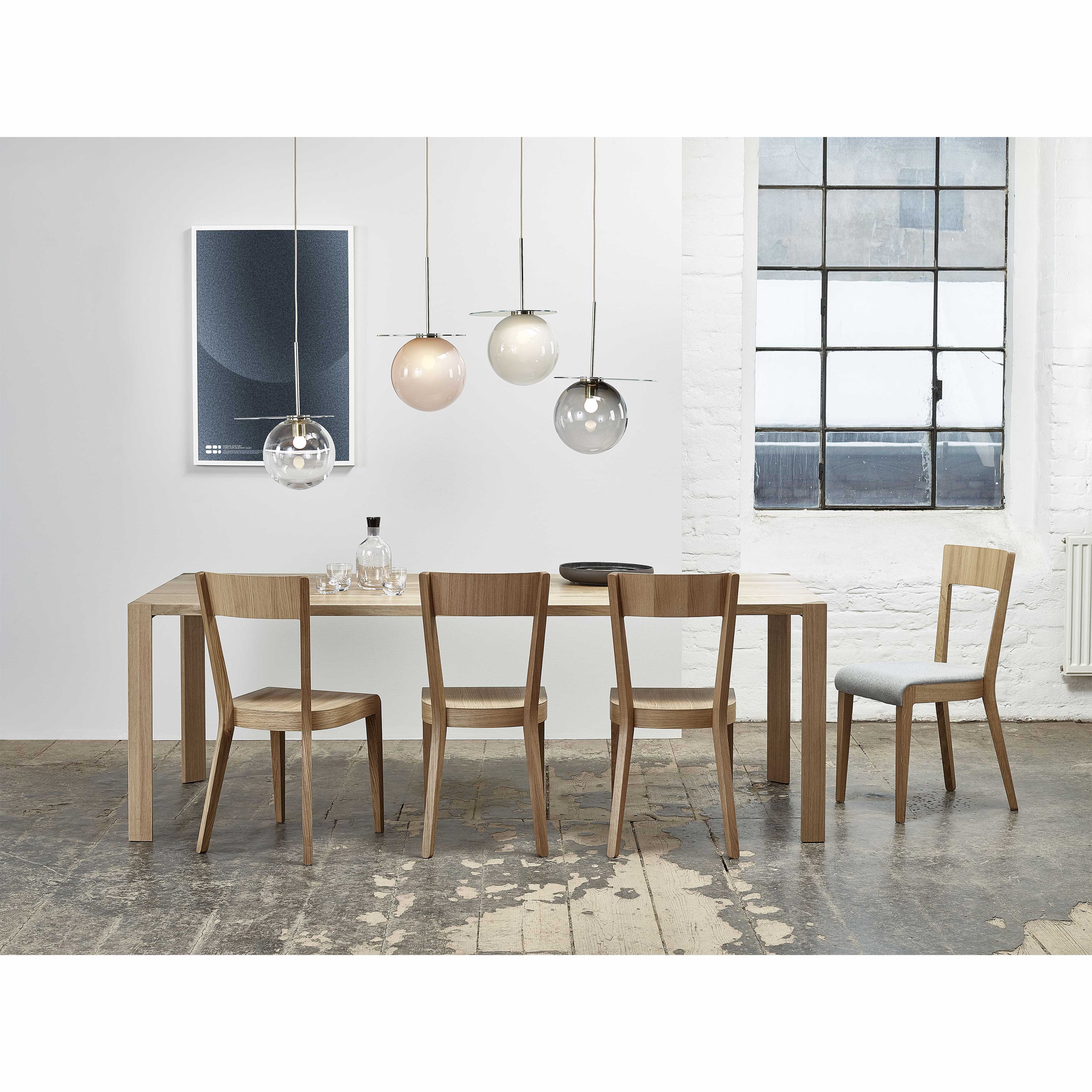A modern dining room showcases a TON Table Ink with triangular legs and six chairs. Four pendant lights hang above the sleek tabletop, while white walls are accented by dark framed windows and minimalist art. A rustic floor adds charm to the space.