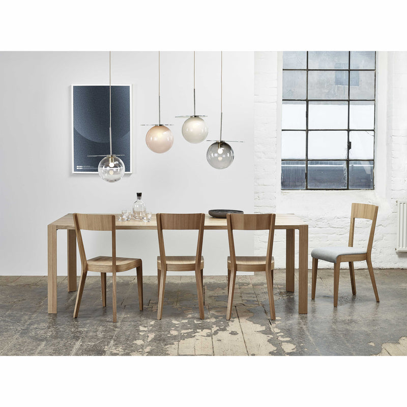 A modern dining room showcases a TON Table Ink with triangular legs and six chairs. Four pendant lights hang above the sleek tabletop, while white walls are accented by dark framed windows and minimalist art. A rustic floor adds charm to the space.