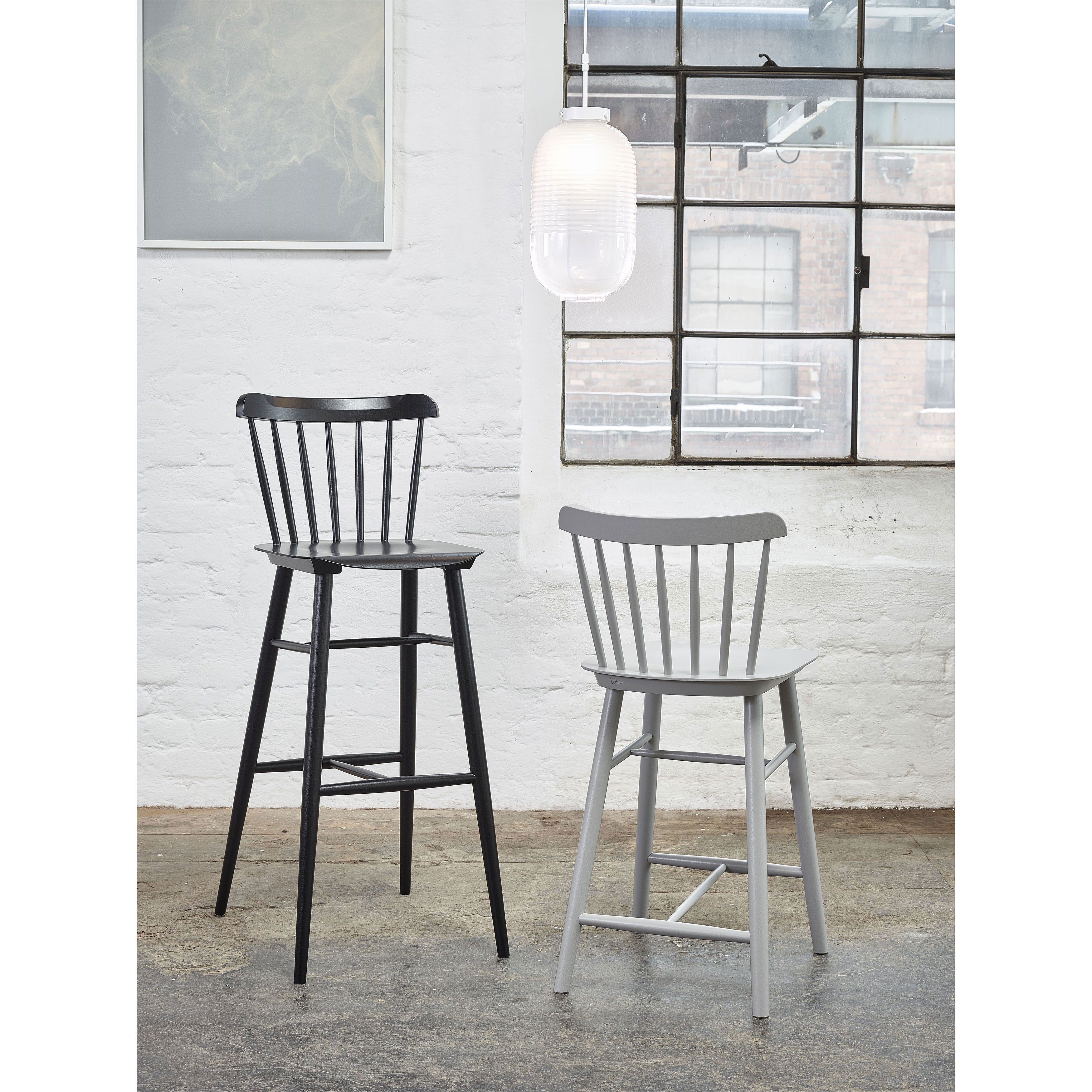 Two Counter Stool Ironica by TON, crafted from beech oak in black and gray, stand on a concrete floor against a white brick wall. A large industrial window offers views of another building, while a modern white pendant light hangs above.