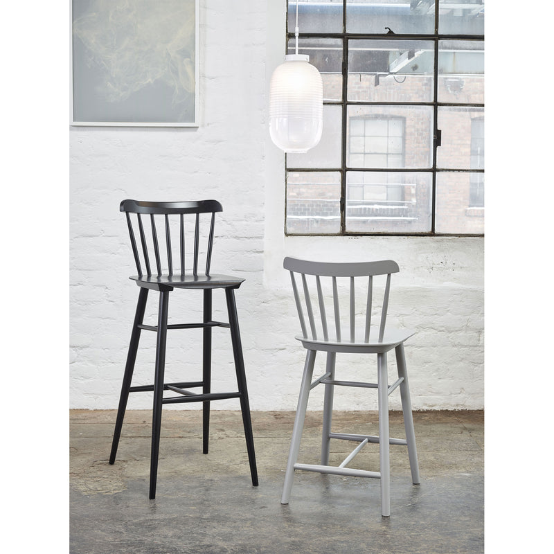 Two Counter Stool Ironica by TON, crafted from beech oak in black and gray, stand on a concrete floor against a white brick wall. A large industrial window offers views of another building, while a modern white pendant light hangs above.