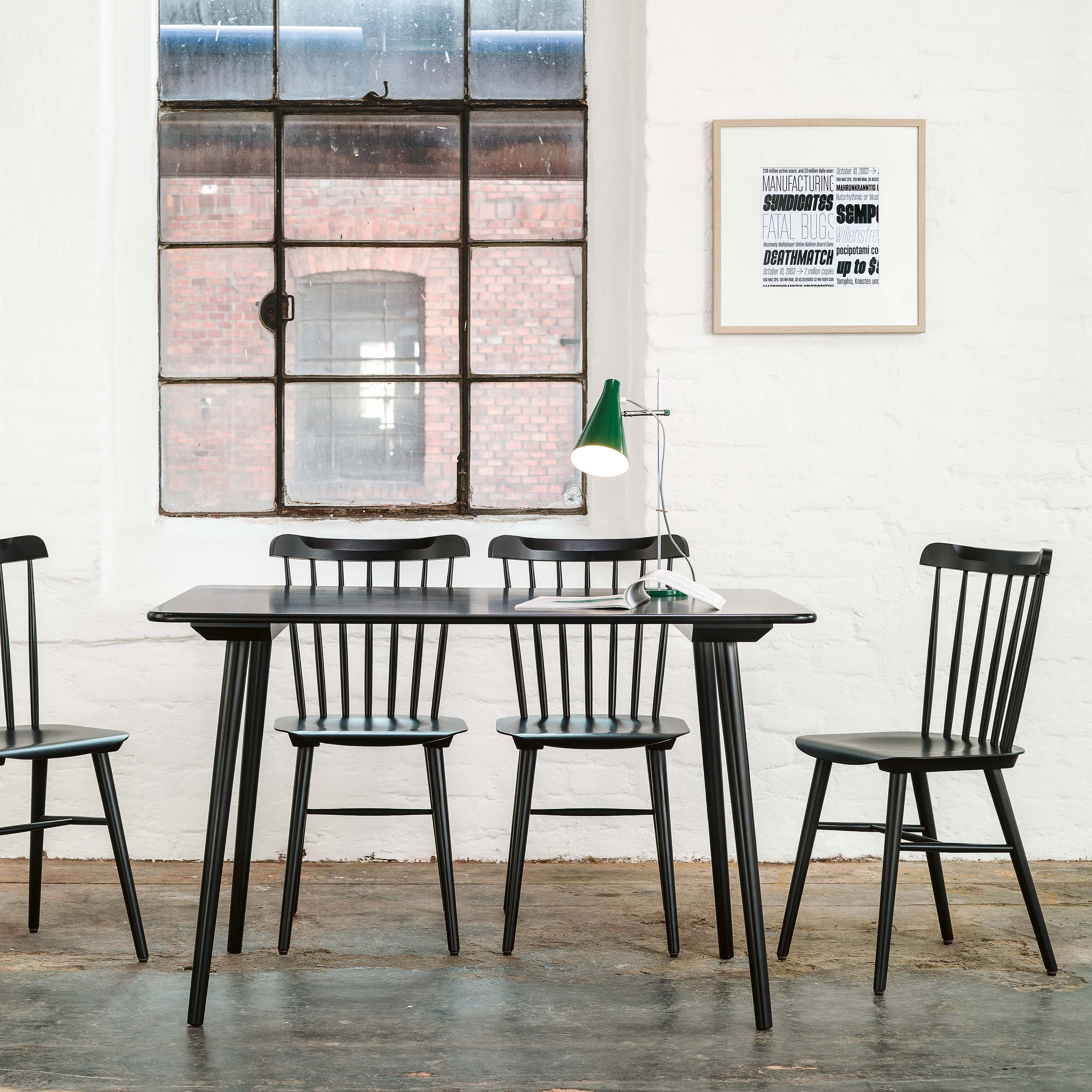 A minimalist dining area includes a rectangular wooden table with four elegant black-spindle TON Chair Ironica - Upholstered chairs. A green desk lamp stands on the table, while a vintage window and framed typographic poster bring retro charm to the white brick wall.