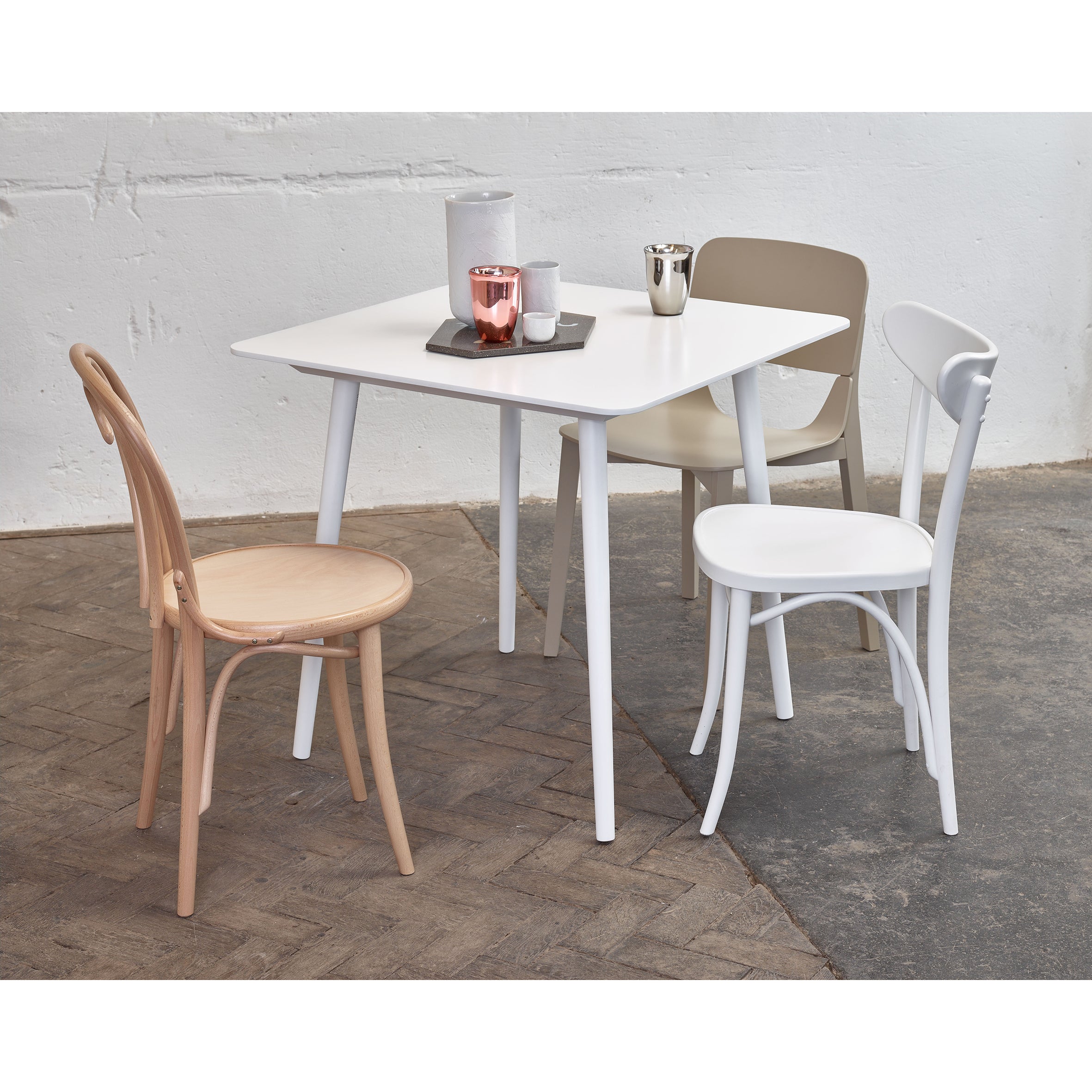 The minimalistic dining setup showcases TON’s Table Ironica, a white square table accented with retro mismatched chairs in white, beige, and solid beech. A gray tray featuring white vases and a copper candle holder perfects the look against a simple white and gray background.